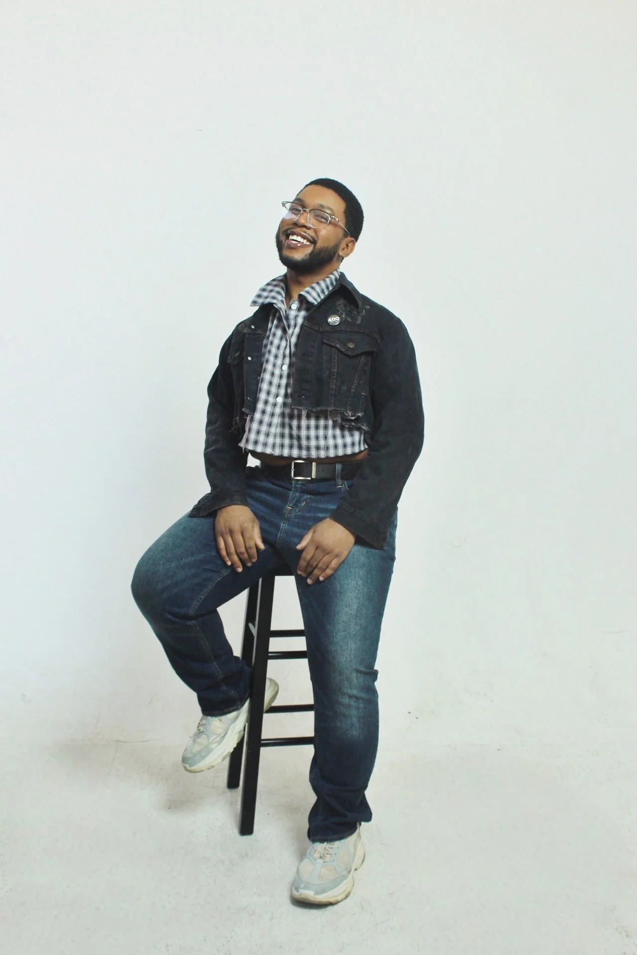 A person with glasses and a beard sitting on a stool, smiling, wearing a black denim jacket, checkered shirt, blue jeans, and white sneakers against a plain white background.