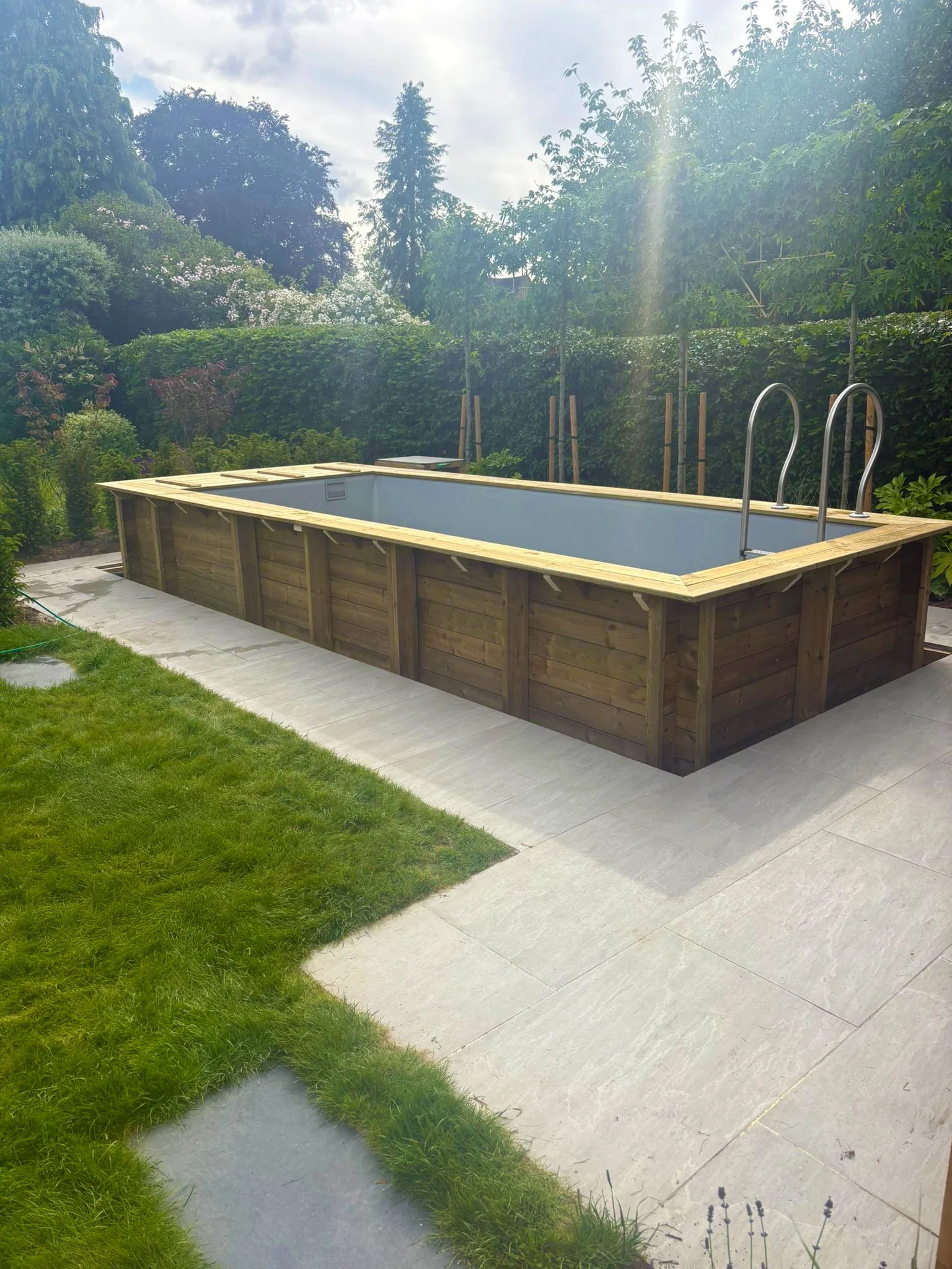 In-ground swimming pool with a wooden deck in a lush backyard with green grass and plants, surrounded by tall trees and hedges.