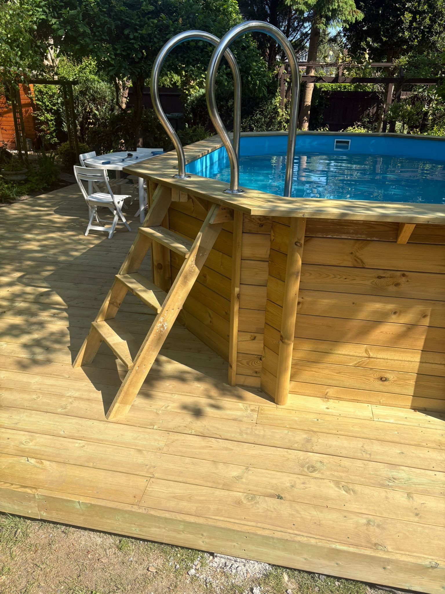 A backyard above-ground swimming pool with a wooden deck and a small ladder, surrounded by trees and outdoor furniture.