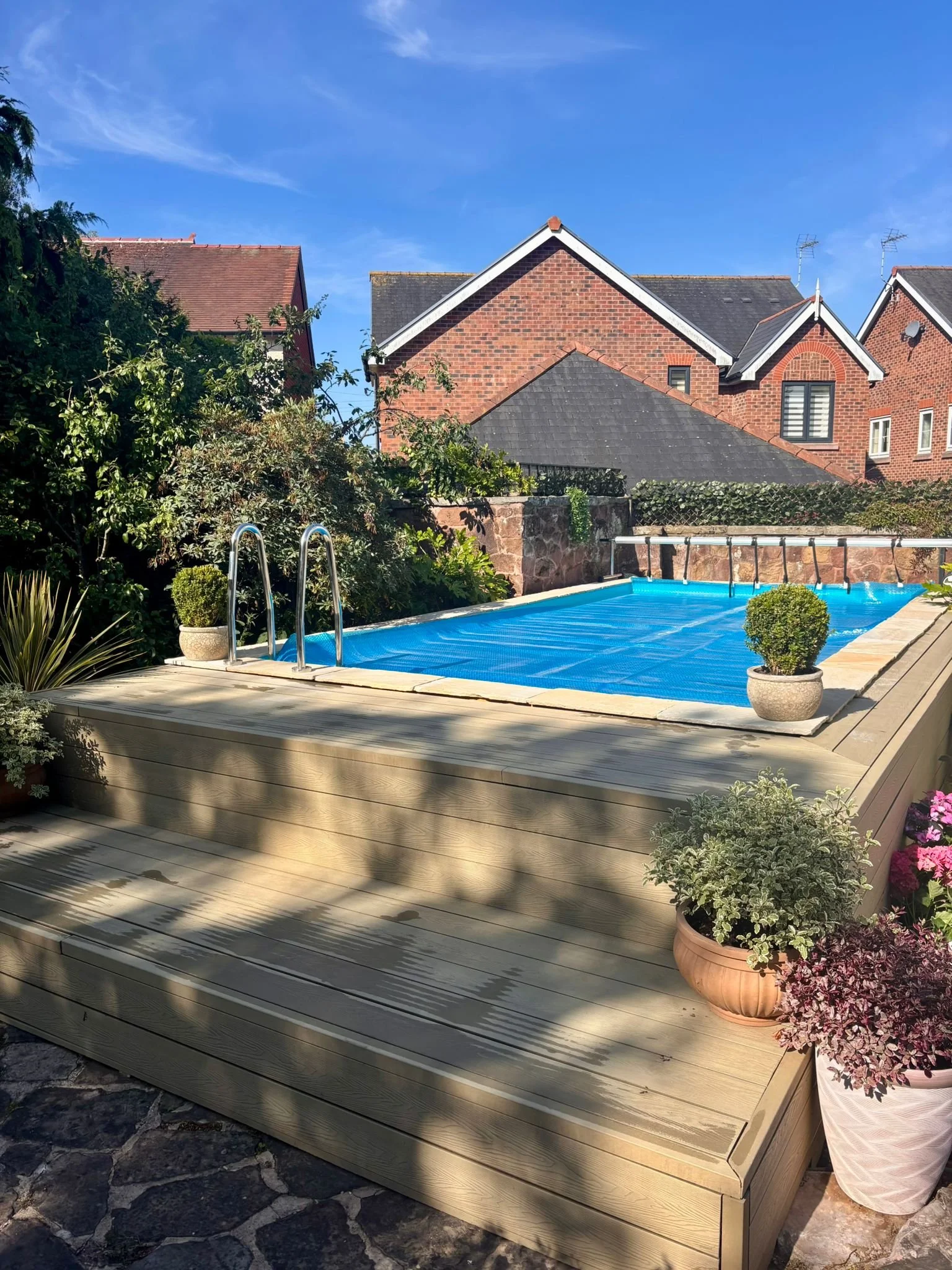 An outdoor swimming pool covered with a blue safety cover, surrounded by potted plants on a wooden deck, with houses in the background under a clear blue sky.