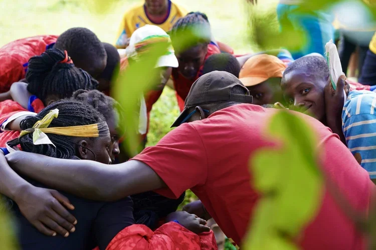 A group of children and a man in red shirt and black cap gathered around outdoors, smiling and interacting closely, with greenery in the background.