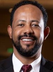 Close-up portrait of a man smiling, wearing a suit and tie, with a blurred dark background.