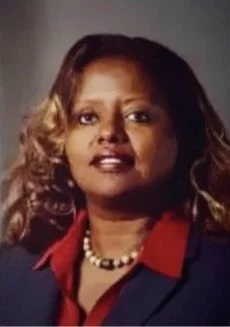 A woman with shoulder-length hair wearing a dark blazer and red shirt with a necklace, posing for a photo.