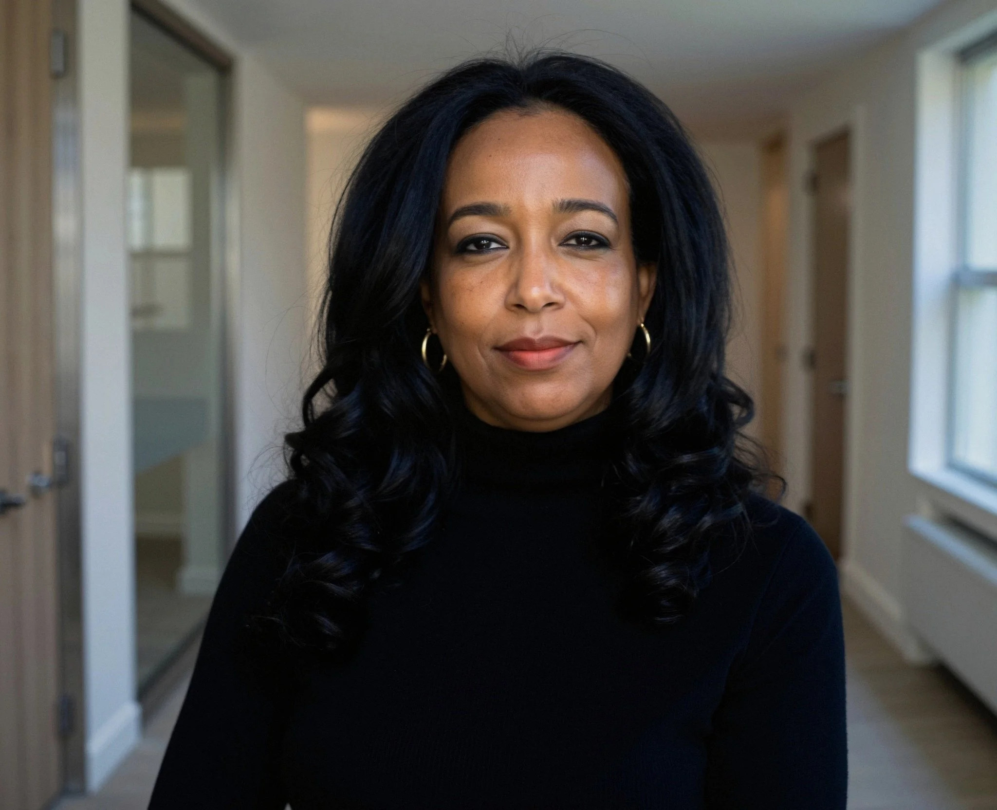 A woman with long, black, curly hair wearing a black turtleneck and gold hoop earrings, standing in a well-lit indoor space.