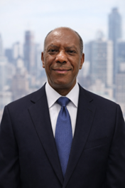 A portrait of an older man in a dark suit and blue tie, standing outdoors with a city skyline in the background.