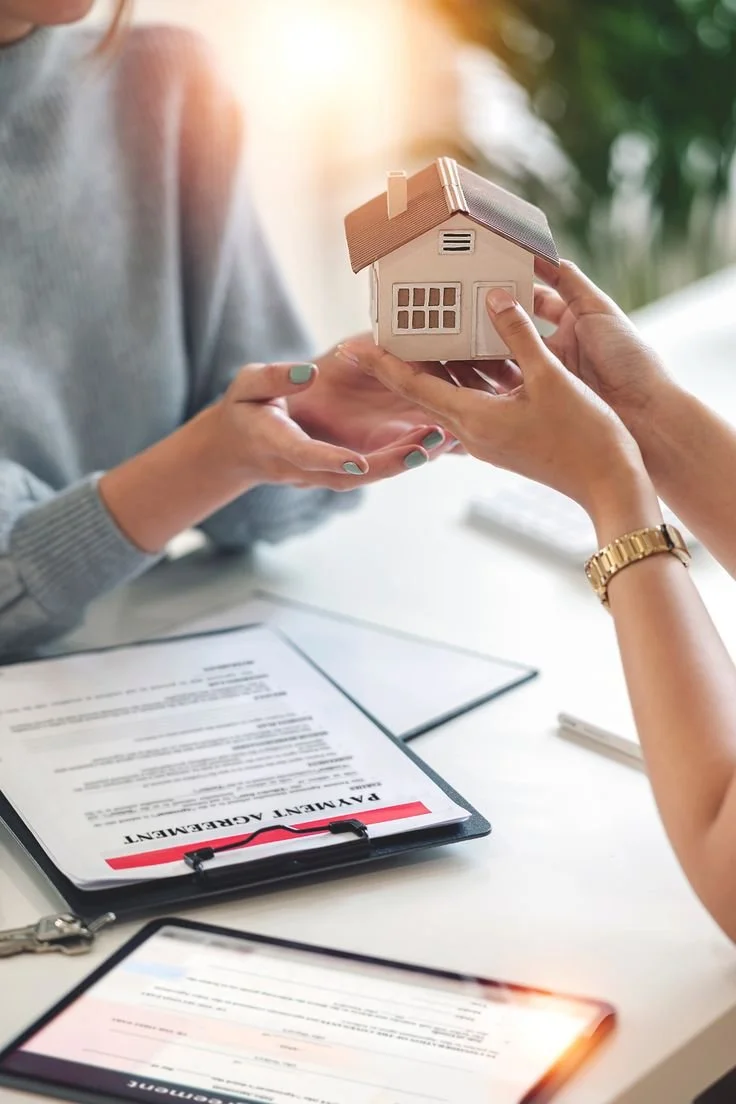 Una mano entrega una maqueta de una casa a otra mano en un entorno de oficina, con documentos y una tableta en la mesa.