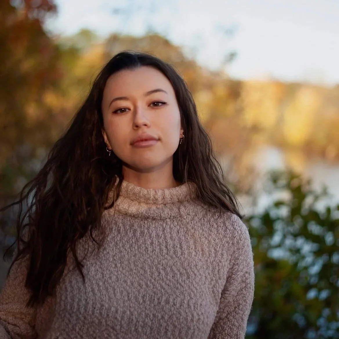 A young woman with long dark hair and hoop earrings standing outdoors in a fall setting with trees and water in the background.