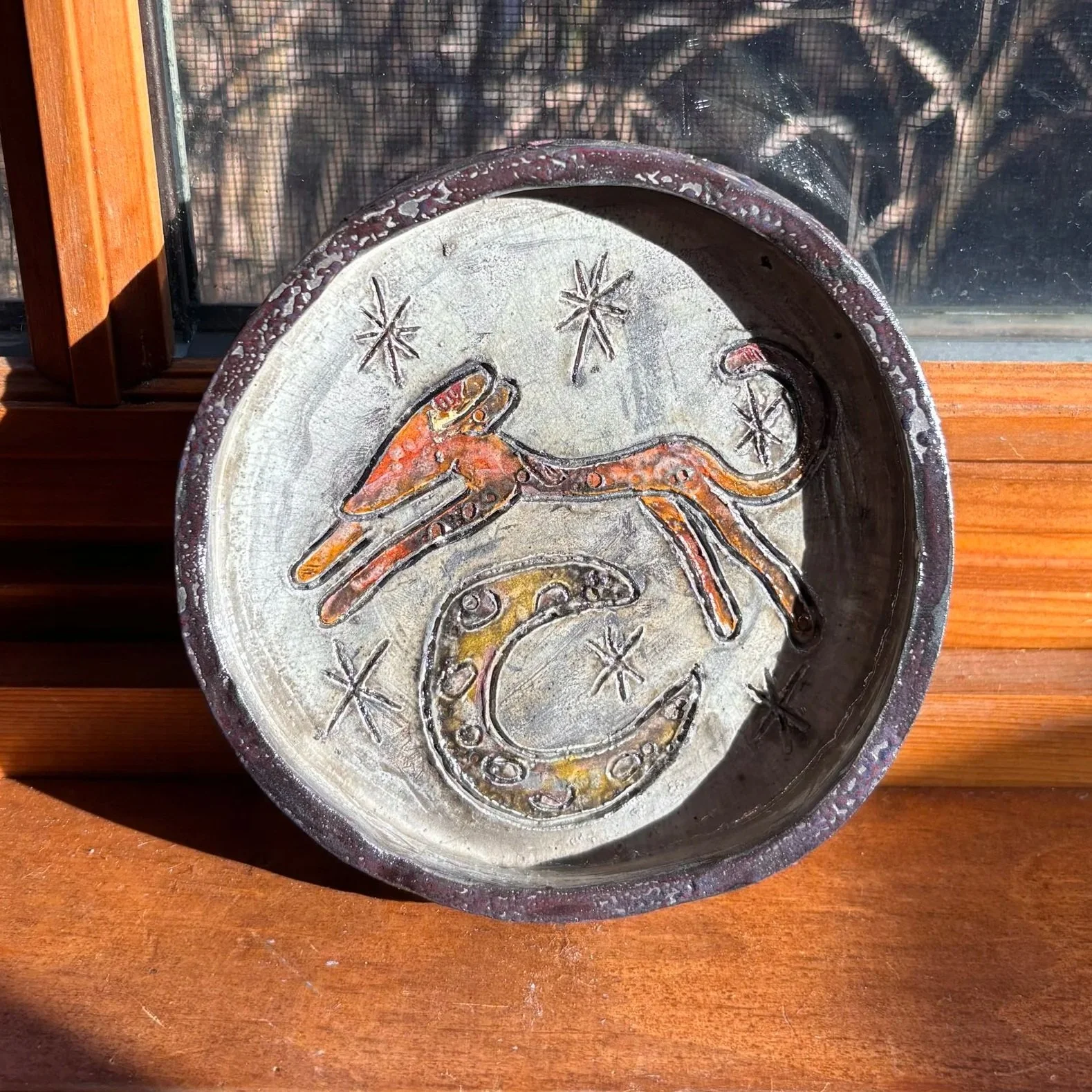 Decorative ceramic dish with a painted octopus, stars, and the letter 'C' on the surface, placed on a wooden ledge.