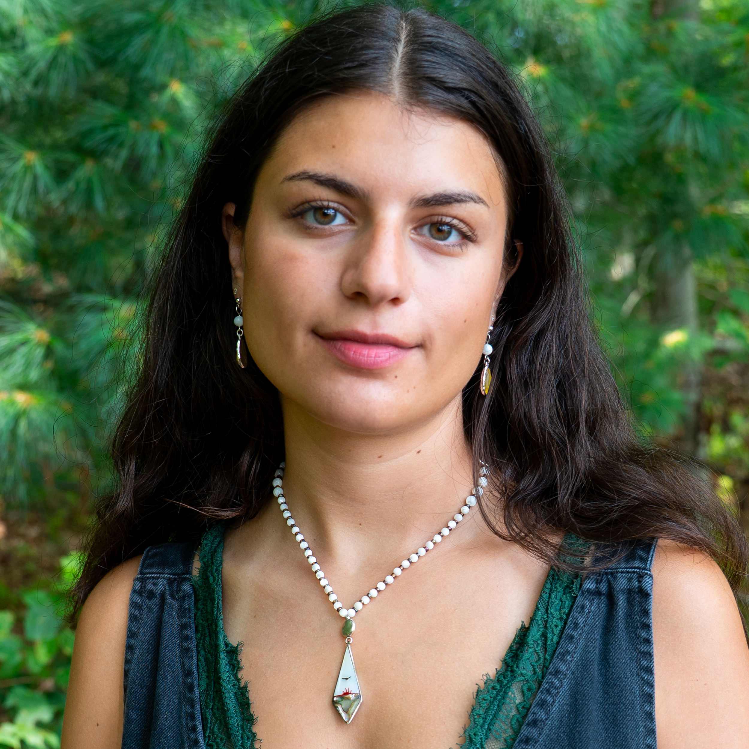 A young woman with dark wavy hair, light skin, and greenish-blue eyes posing outdoors in front of green foliage, wearing earrings, a pearl necklace with a pendant, and a denim sleeveless top with lace detail.