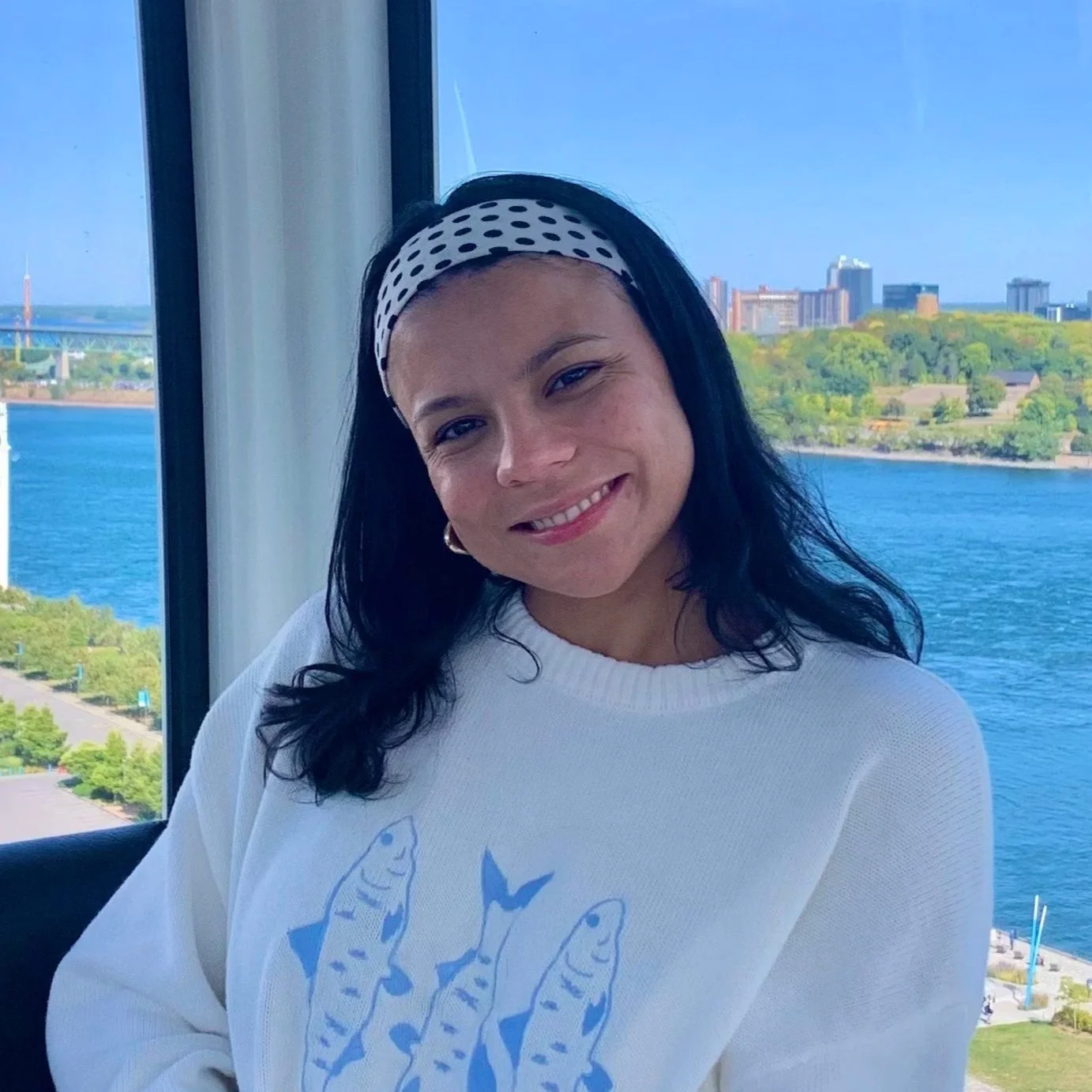 A woman with long dark hair wearing a white sweater with blue fish design and a polka-dot headband, smiling in front of a large window overlooking a river, cityscape, and green trees.