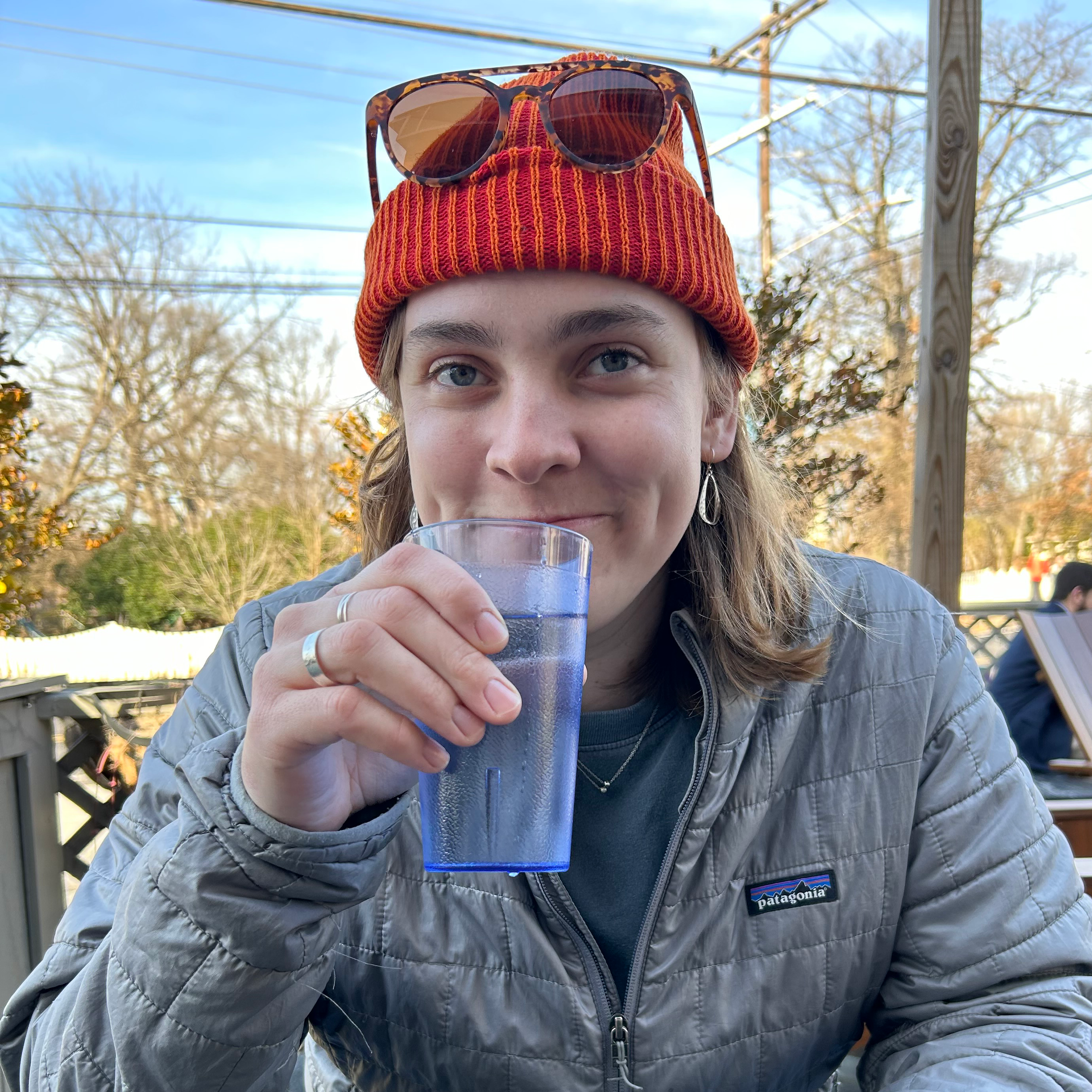 Young woman wearing a red beanie with sunglasses on top, drinking water from a blue glass outdoors, with trees and a wooden fence in the background.