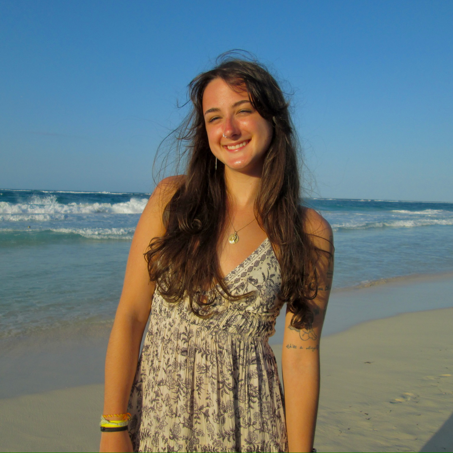 A smiling young woman with long dark hair standing on a beach with the ocean and waves in the background.
