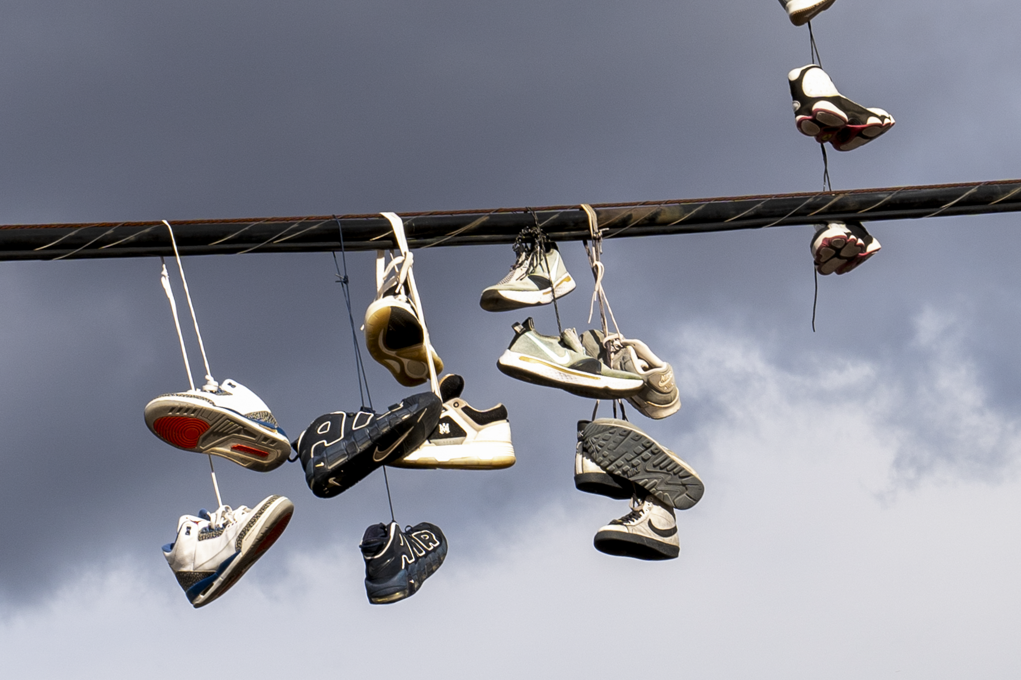 Multiple sneakers hanging from a wire against a cloudy sky.