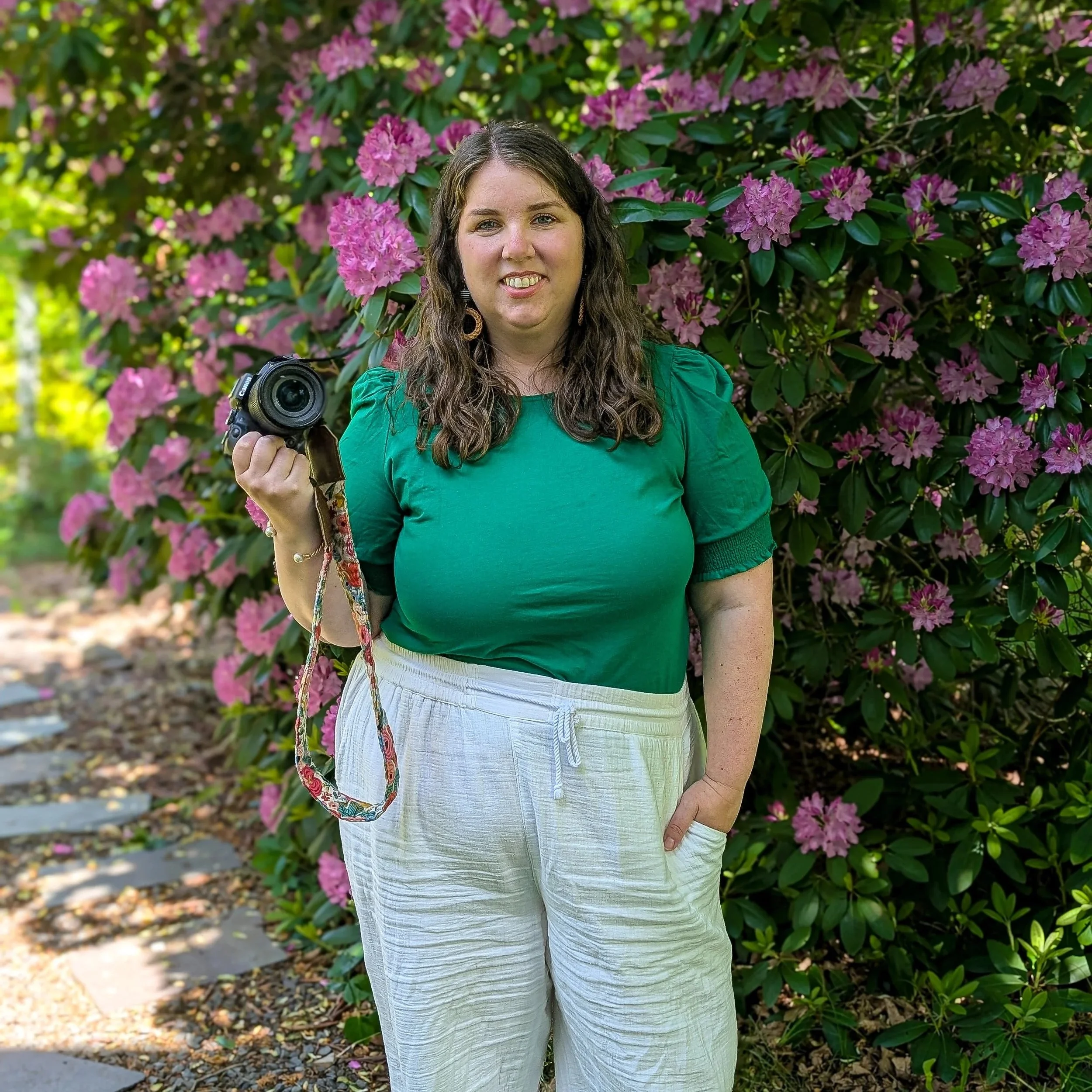 A woman with brown curly hair, wearing a green top and white pants, holding a camera, standing in front of a flowering bush with pink flowers.