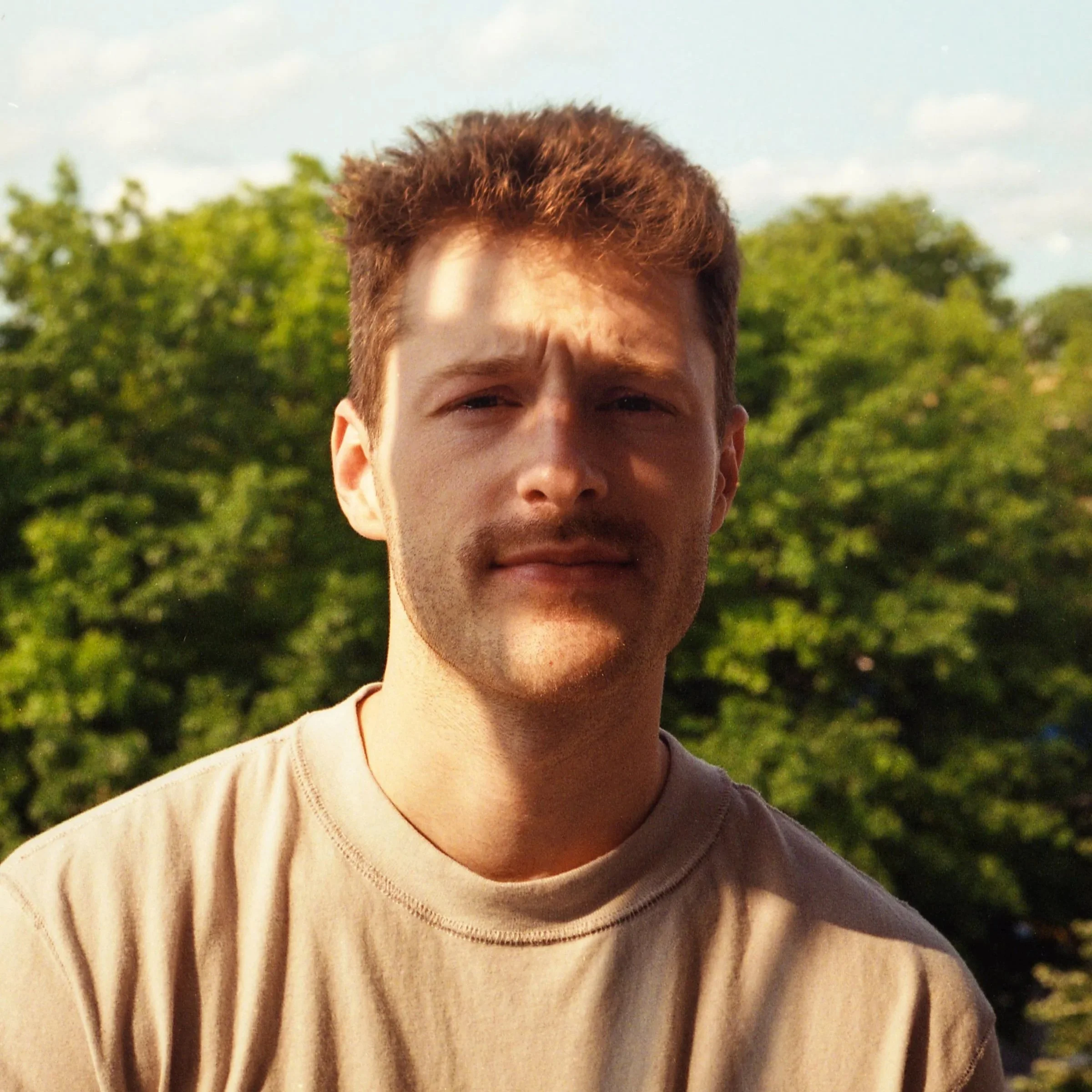 A young man with light skin, short brown hair, and facial hair stands outdoors with trees and a partly cloudy sky in the background.