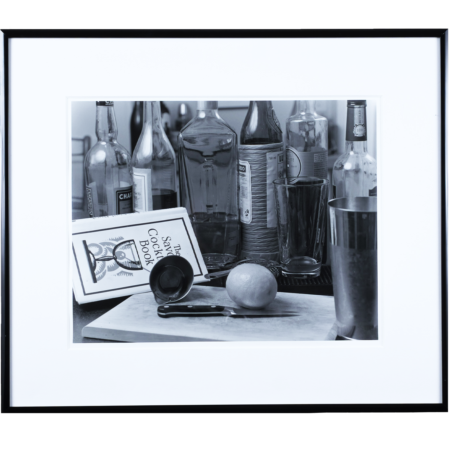 Black and white photo of various bottles, glasses, a knife, an apple, and a cocktail recipe book on a table.