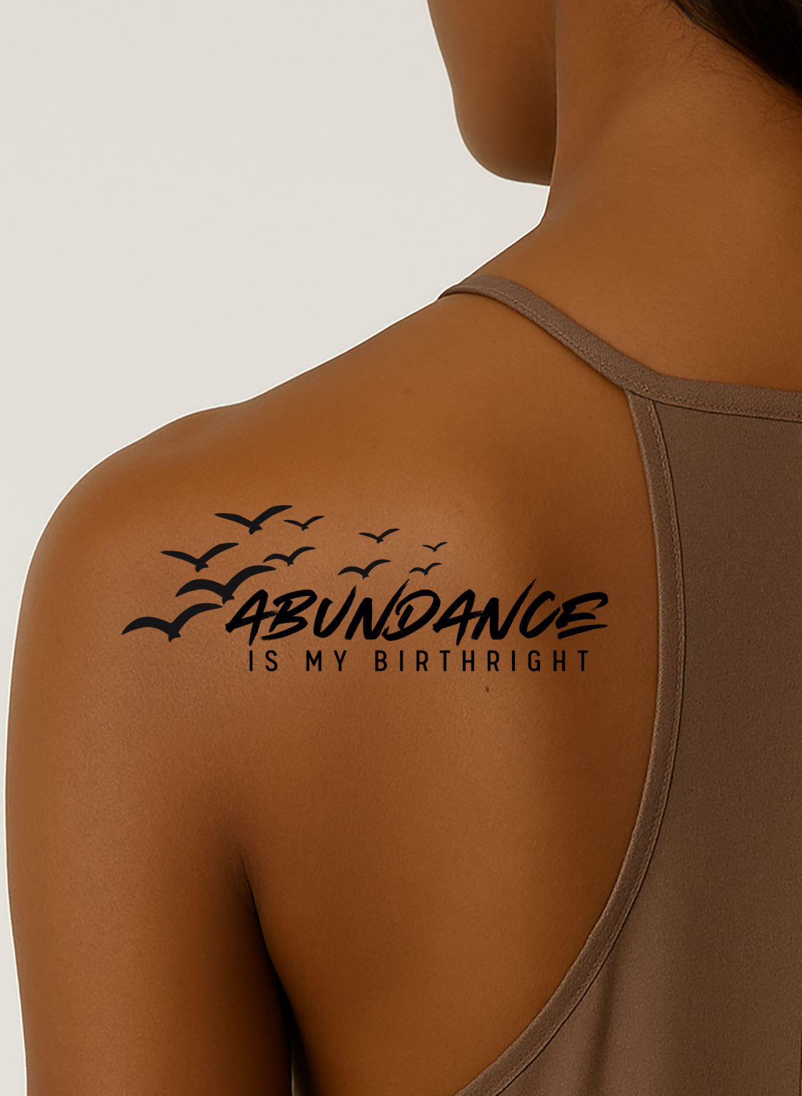 Female with brown skin color has a tattoo on her left shoulder that reads "Abundance is my birthright" and it has seagulls flying around it.