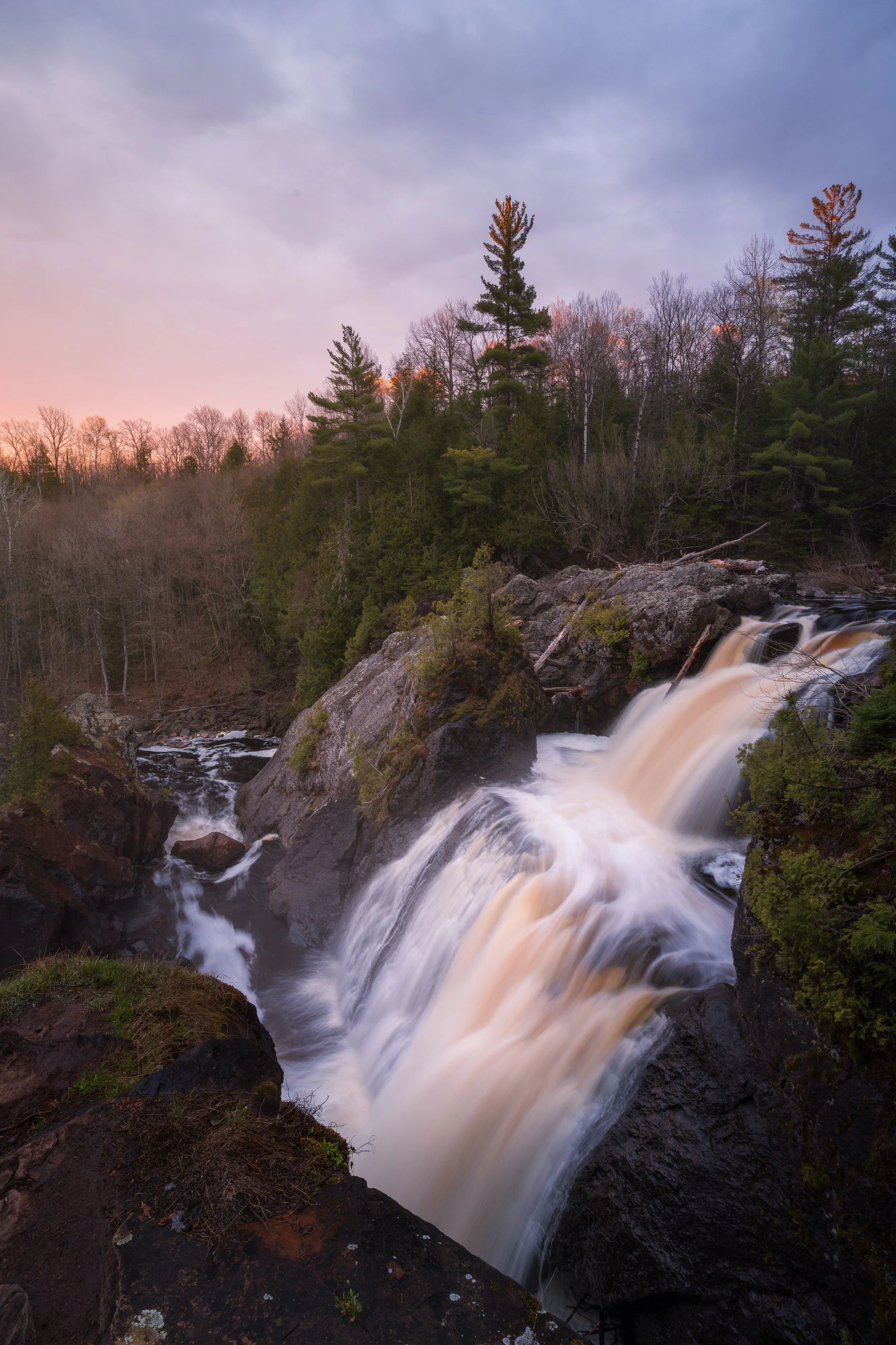 A waterfall flowing over rocks in a forest with trees, some without leaves, during sunset or sunrise with a cloudy sky.