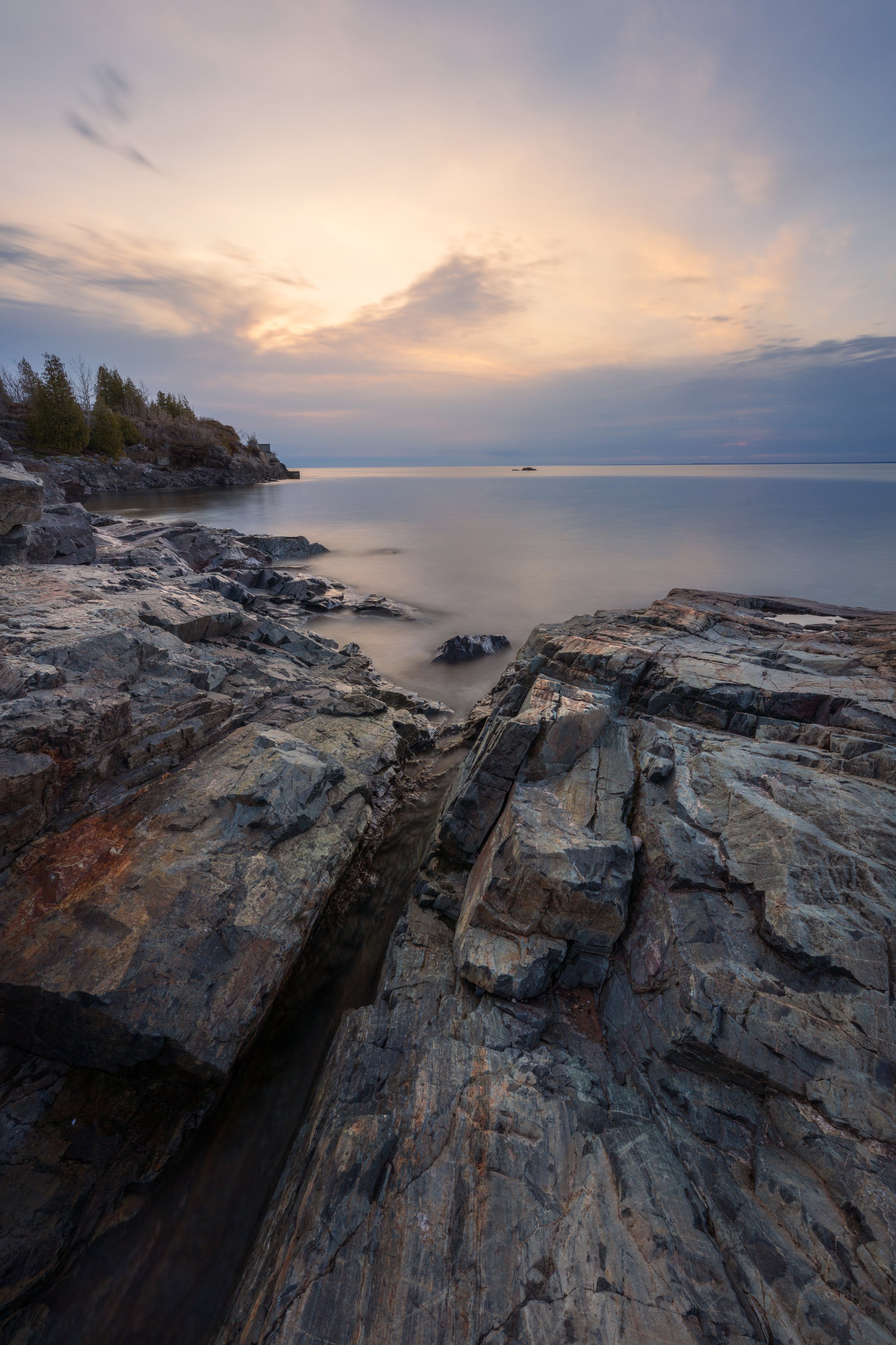 A rocky shoreline with large, layered rocks in the foreground and calm water extending to the horizon. The sky above is filled with soft clouds, with the sun either setting or rising, creating a peaceful, slightly overcast atmosphere.
