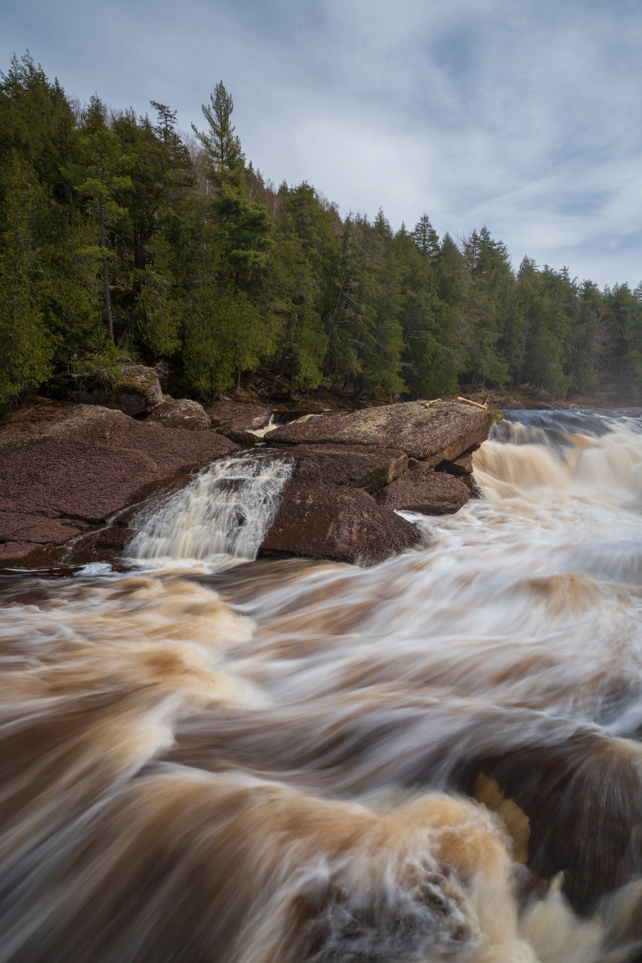 A river flowing over rocks with a forested hillside and cloudy sky in the background.