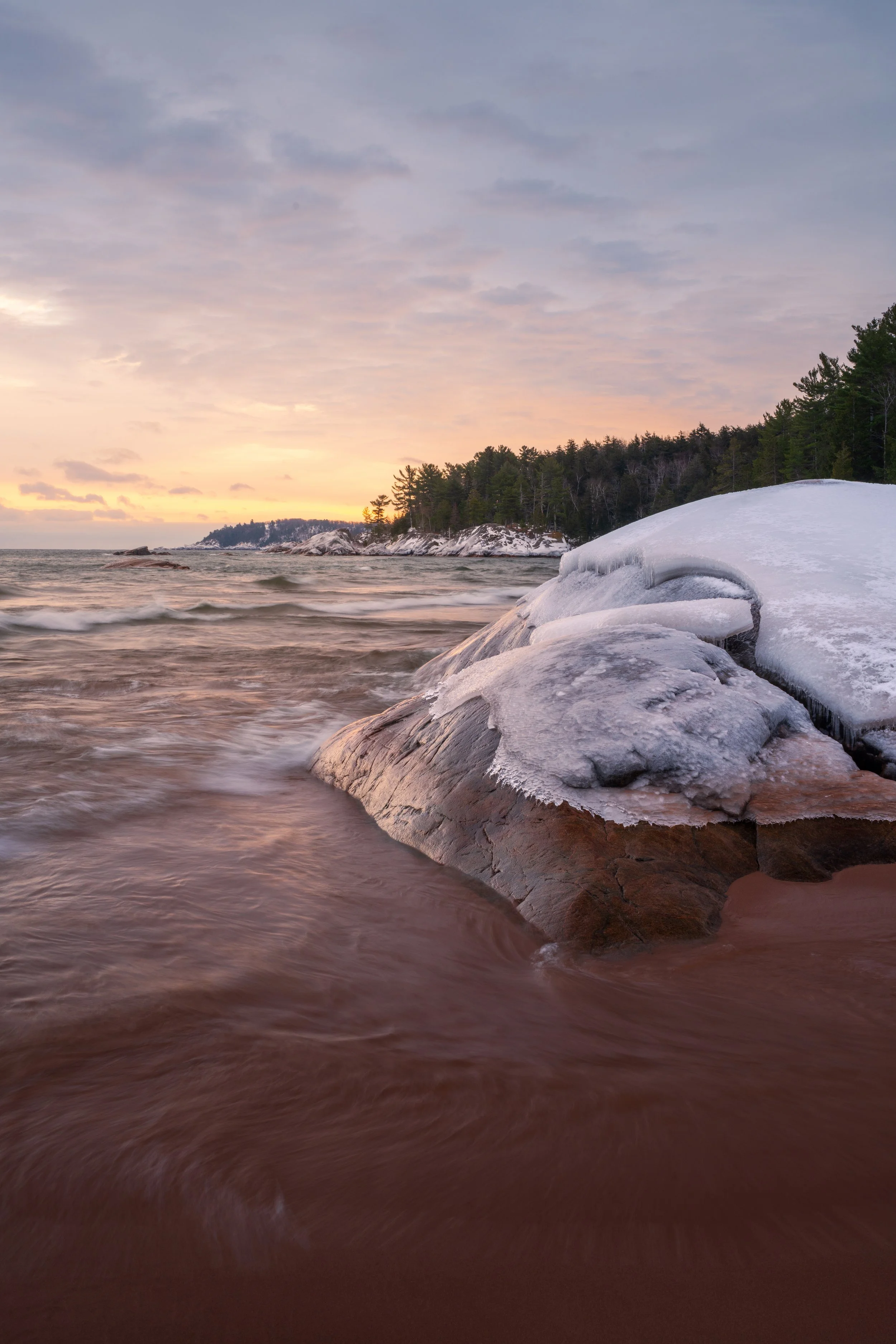 A beach at sunset with a rocky shoreline and trees in the background, with snow or ice on the rocks in the foreground.