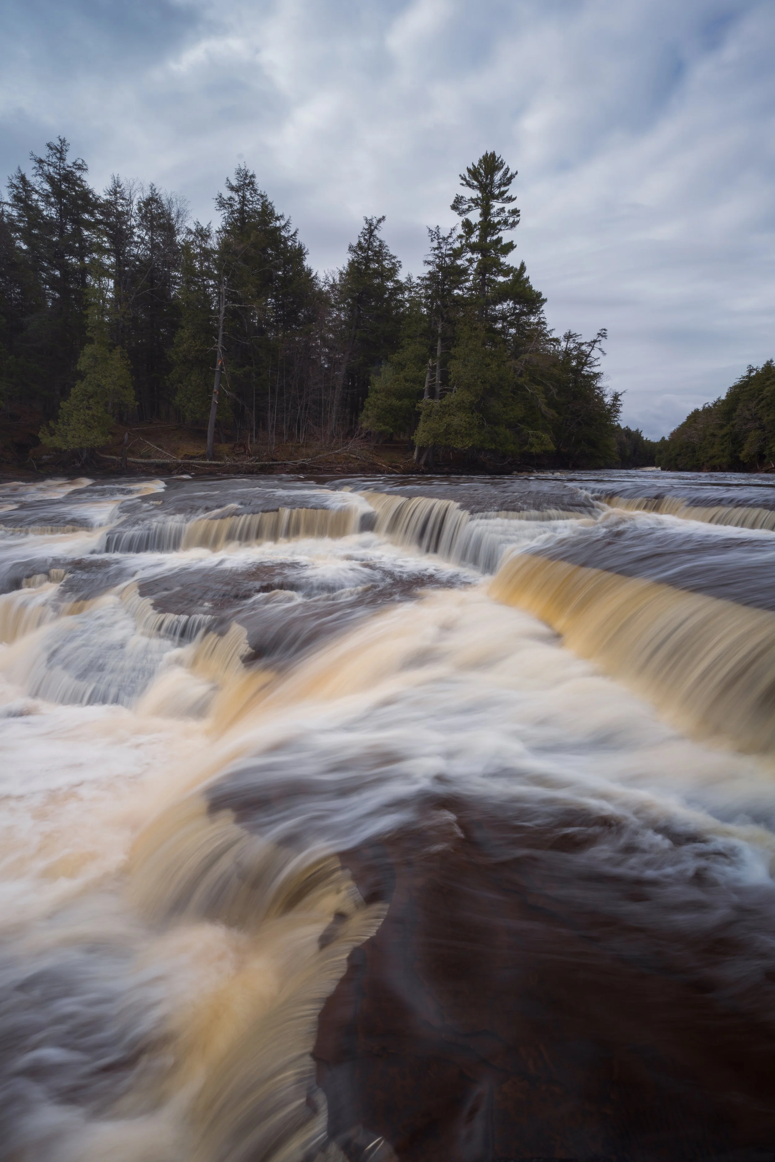 A wide river with rapids flowing through a forested area under a cloudy sky.