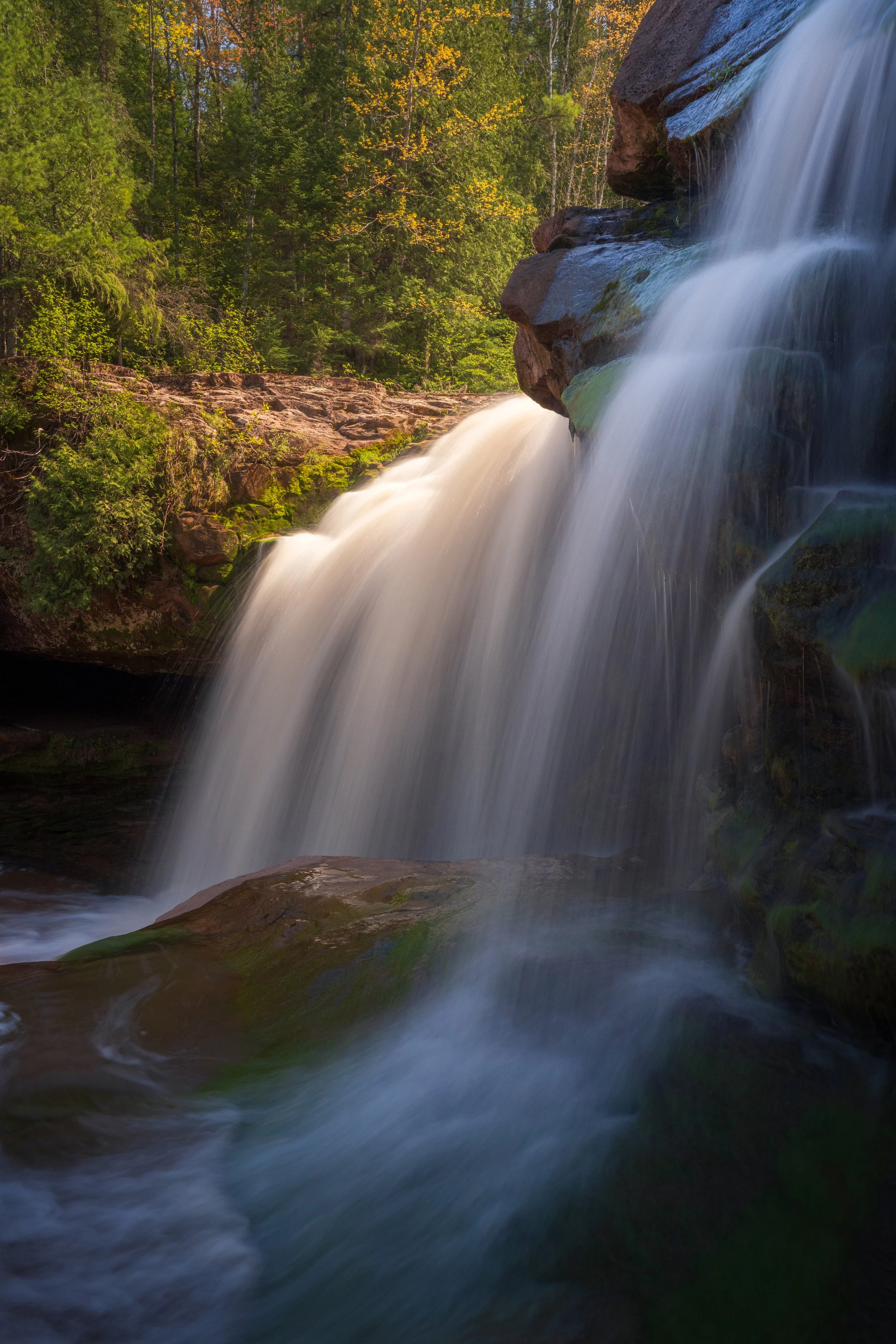 A waterfall cascading over rocks in a forest setting with lush green trees and some yellow leaves in the background, sunlight illuminating parts of the scene.
