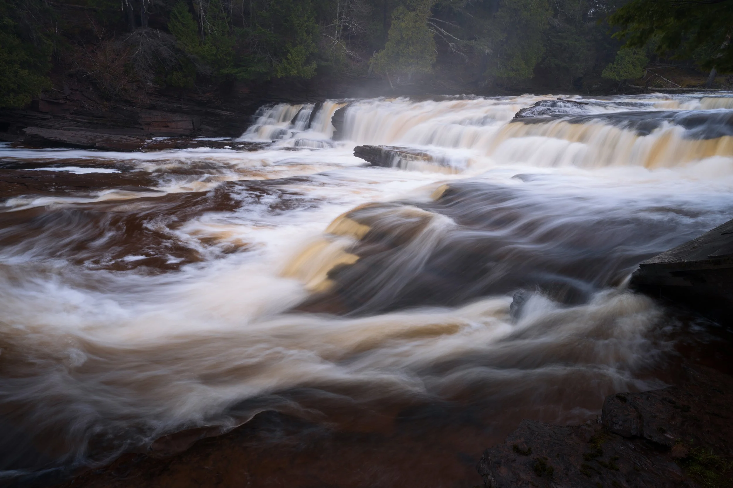 A scenic view of a waterfall flowing over rocks with trees along the riverbank in the background.