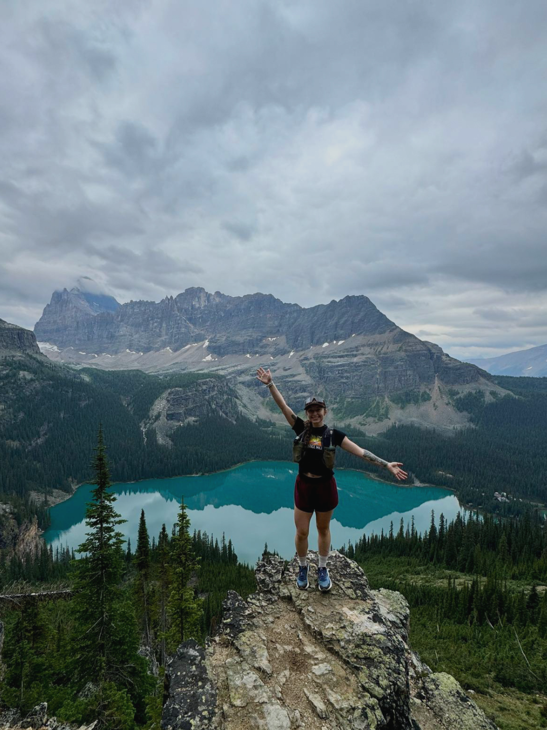 Smiling woman standing on a rock with arms open, overlooking a mountain lake surrounded by trees and mountains on a cloudy day.