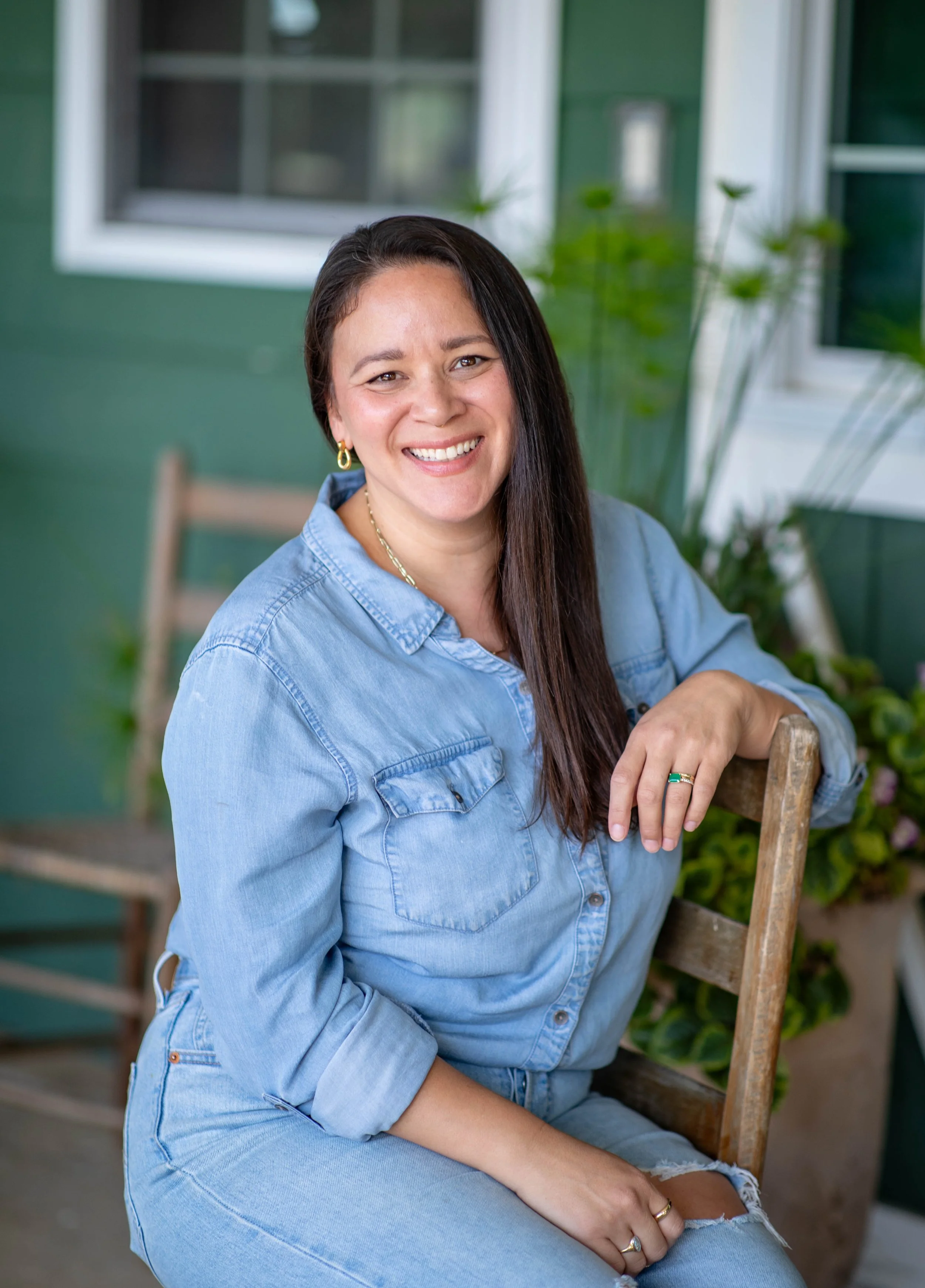 Smiling woman with long dark hair wearing a light blue denim shirt sitting outdoors on a wooden chair next to green house siding, plants, and a window.