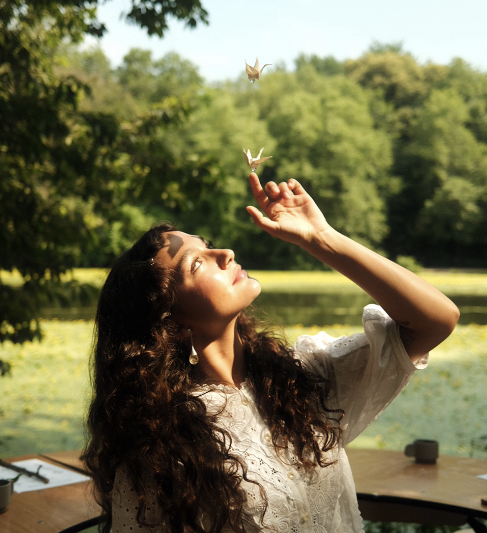 A woman with long curly hair is sitting outdoors near a lake with green trees in the background. She is raising her hand and appears to be releasing or watching two paper origami cranes flying above her, illuminated by sunlight.