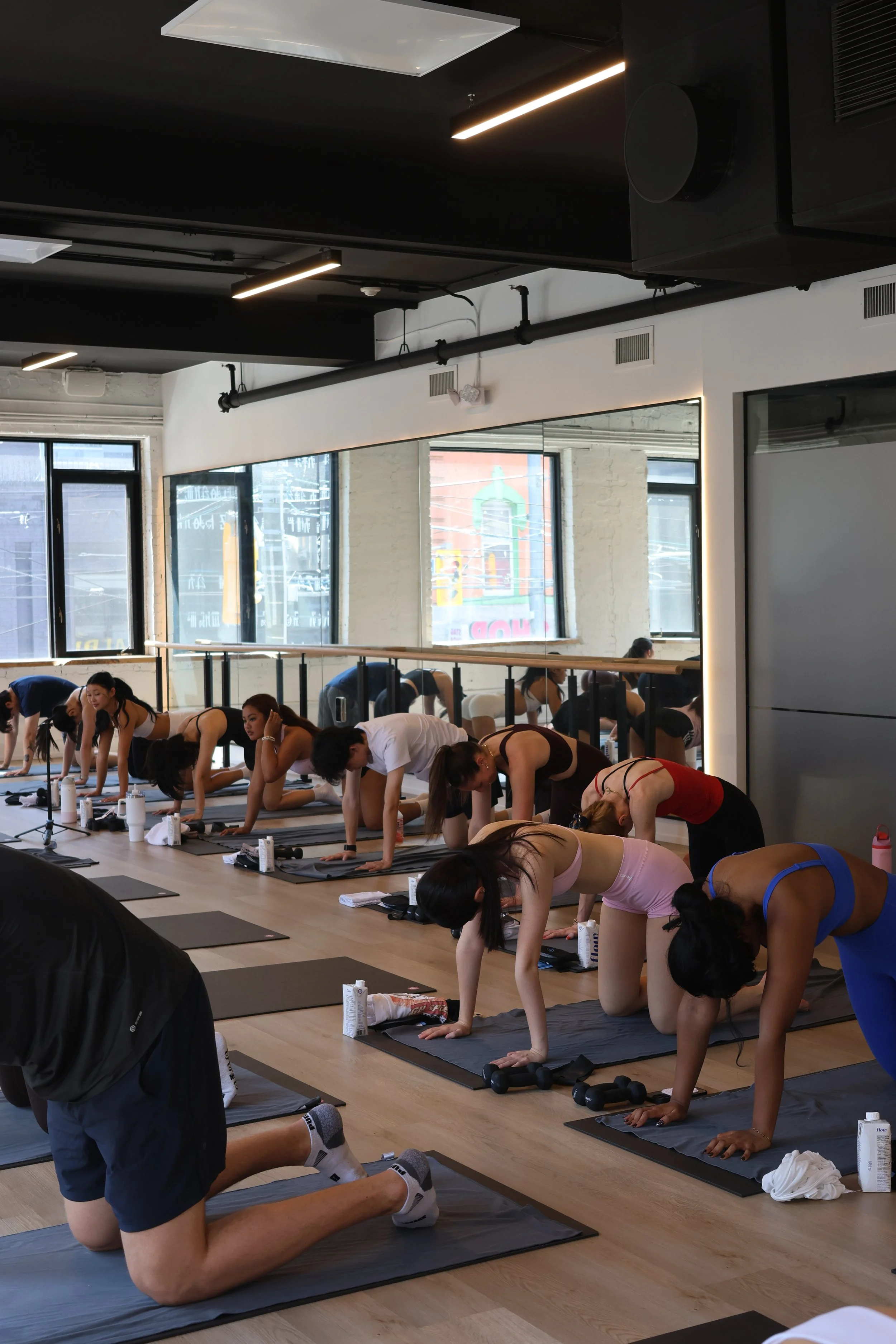 A group of people participating in a yoga or Pilates class in a studio with large windows, black and white walls, and modern lighting fixtures.