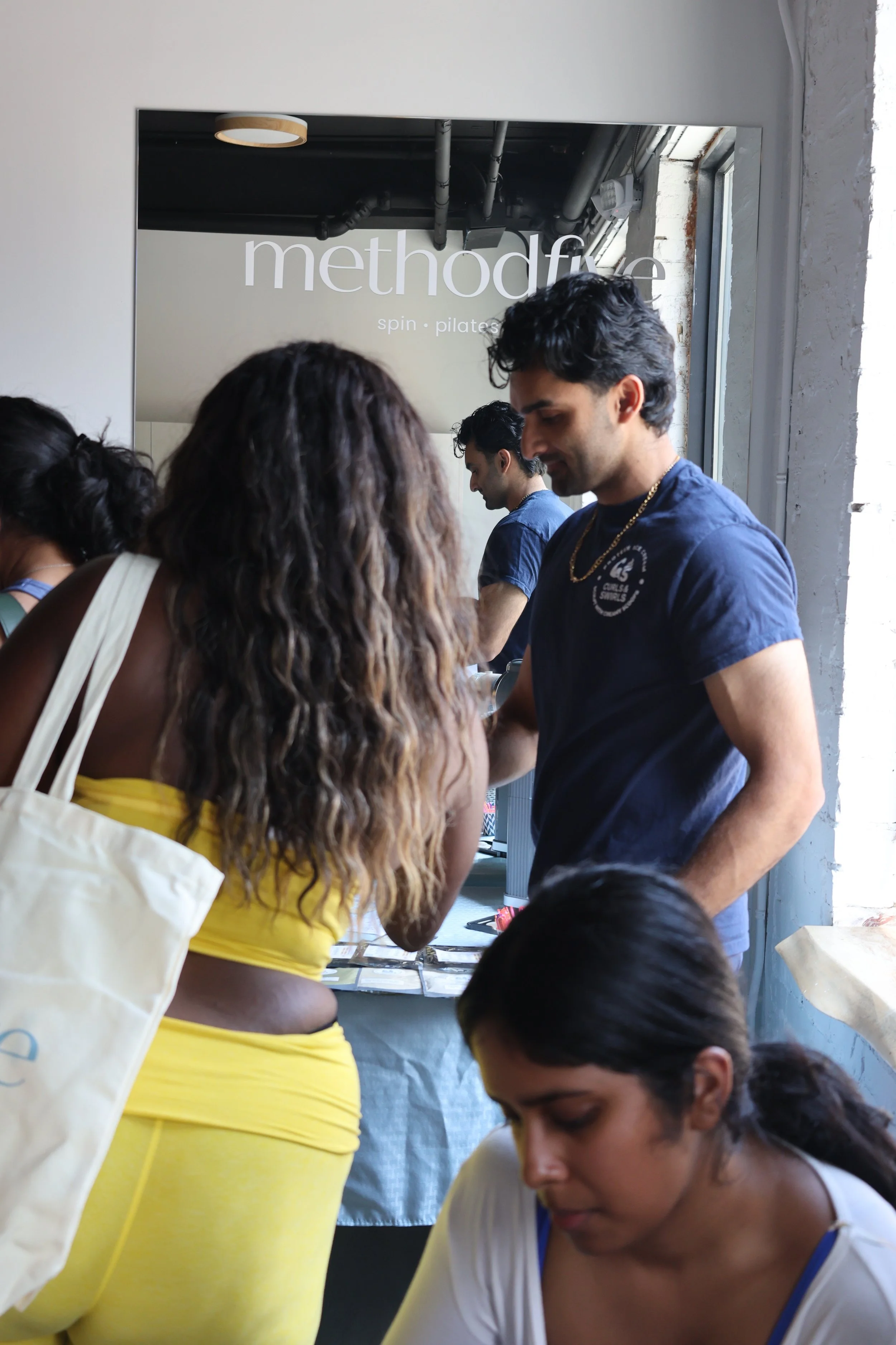 People gathered around a table inside a studio named 'methodfy' with a sign for spin and pilates classes, in an industrial-style space with exposed pipes and a window.