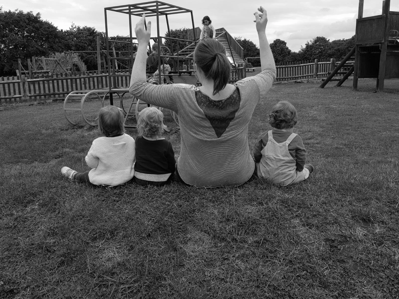 A woman sitting on the grass with three children, facing a playground with a child climbing a structure. The woman has her arms raised, and the scene is outdoors on a cloudy day.