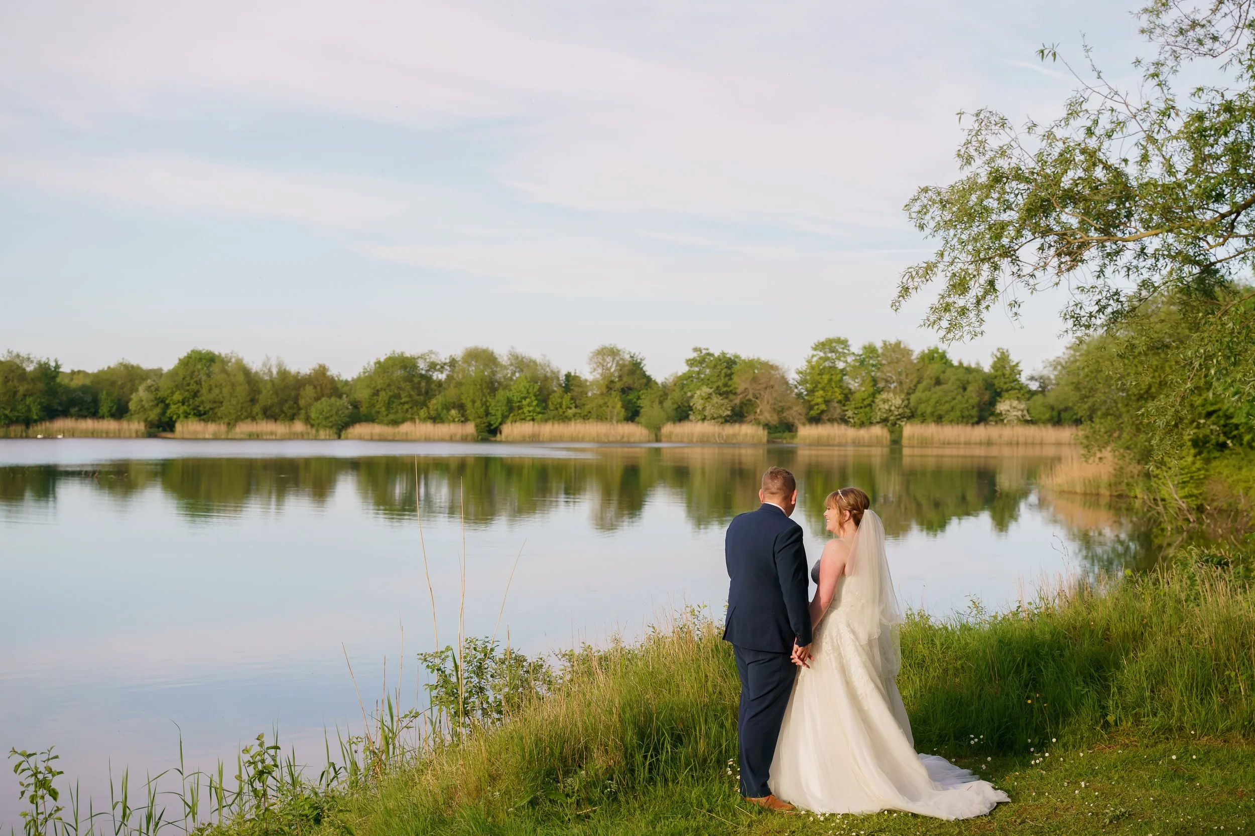 A bride and groom standing hand in hand near a lake, surrounded by green trees and grass, with a cloudy sky in the background.