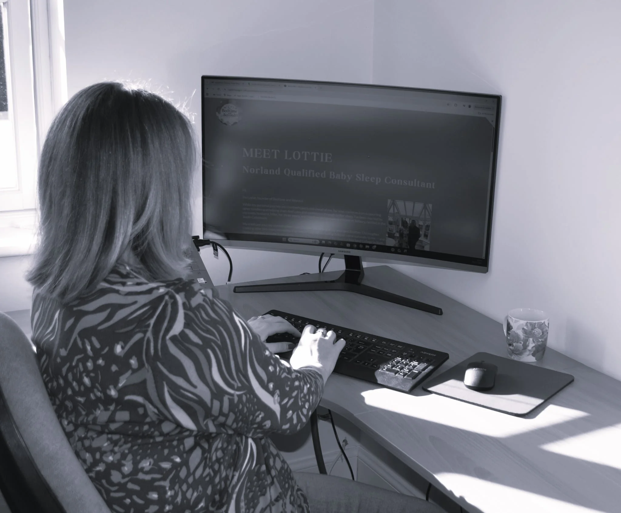 A woman sitting at a computer desk, viewing a website about a baby sleep consultant, with a monitor, keyboard, mouse, a cup, and a notebook on the desk.