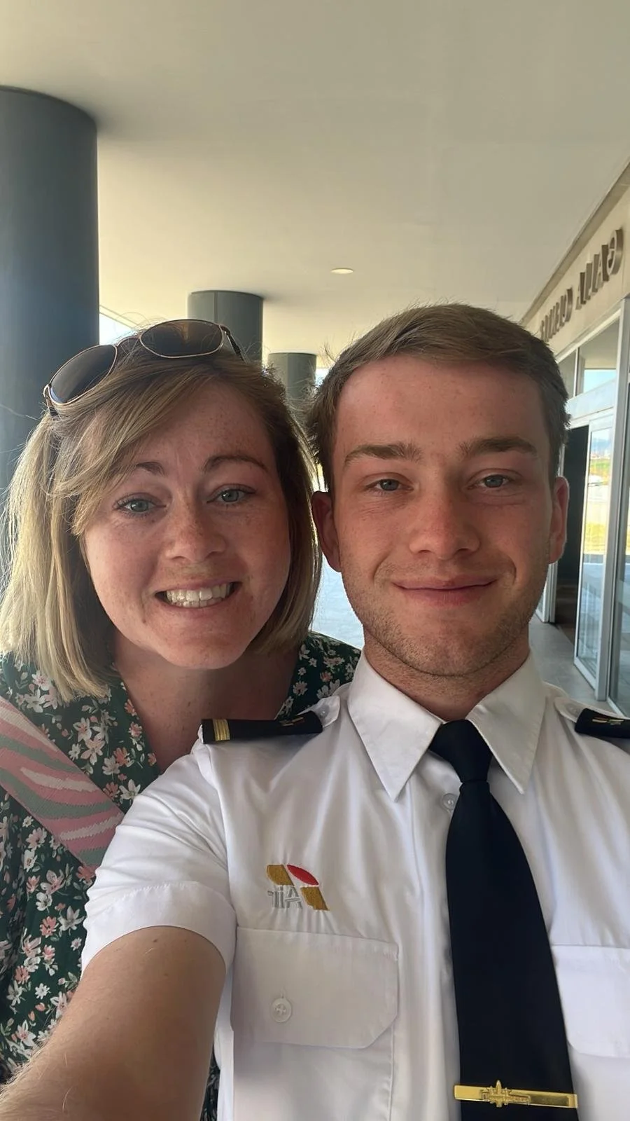 A woman and a man taking a close-up selfie together. The woman has light brown hair, sunglasses on her head, and is smiling. The man is dressed in a pilot's uniform with a white shirt, black tie, and epaulets, also smiling. They are outside a building with large windows.