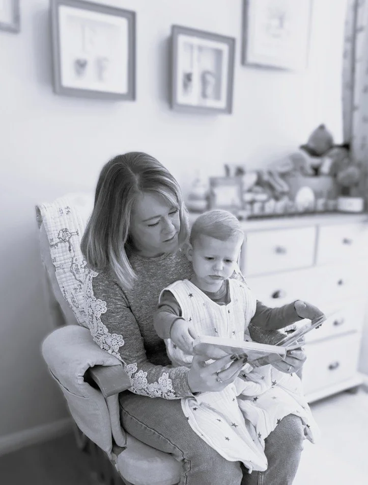 A woman and a young girl reading a book together while sitting on a cushioned armchair in a cozy room with framed pictures on the wall and drawers in the background.