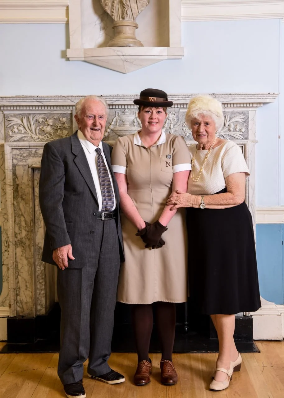 Three people standing in front of a decorative fireplace, holding hands, smiling. An older man in a gray suit and a woman in a beige dress and bowler hat in the center, holding hands with an elderly woman in a white blouse and black skirt on the right.
