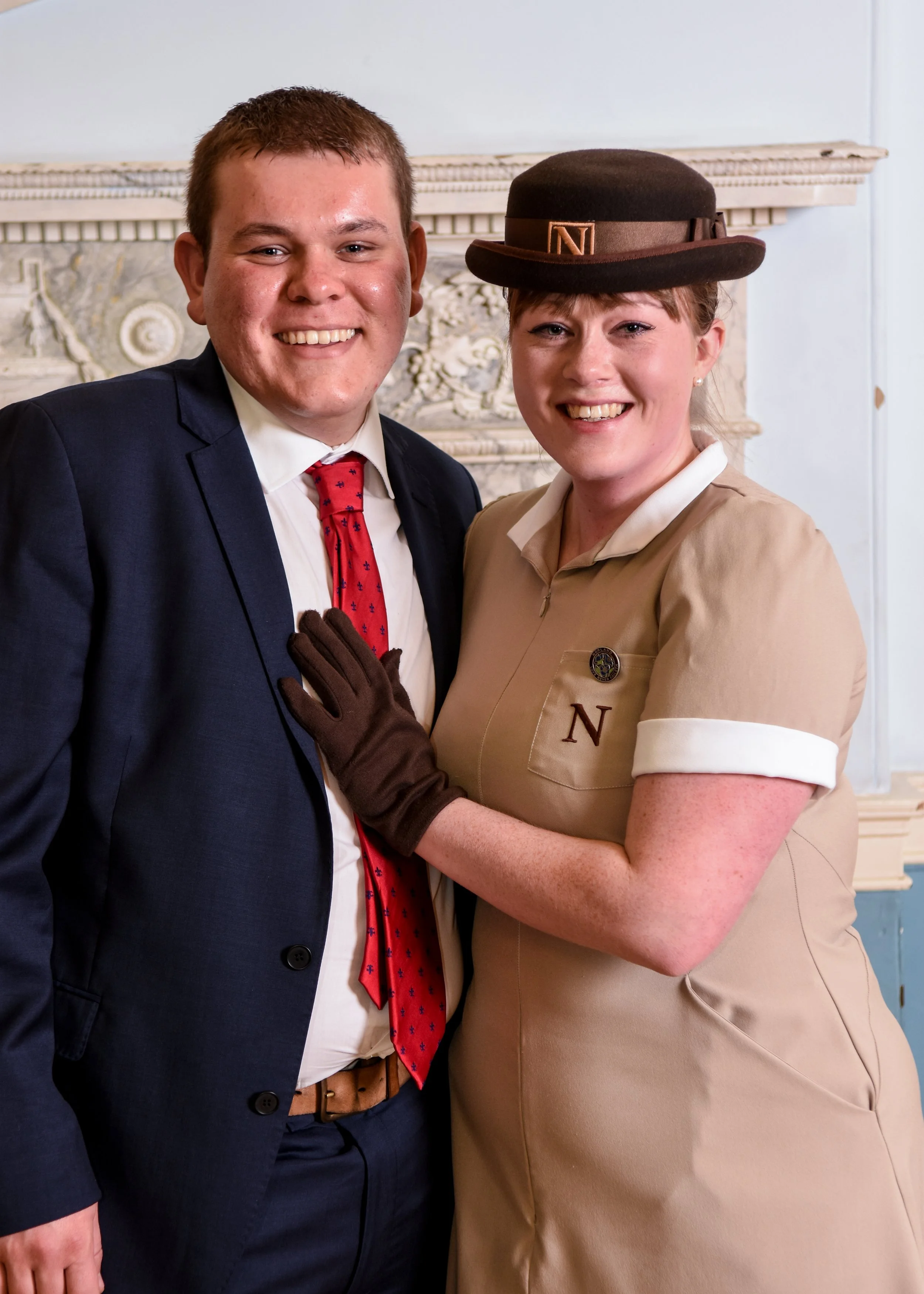 A man and a woman smiling and posing together, the woman wearing a vintage-style uniform with an 'N' badge and a hat, and the man in a suit with a red tie, standing in front of an ornate stone fireplace.