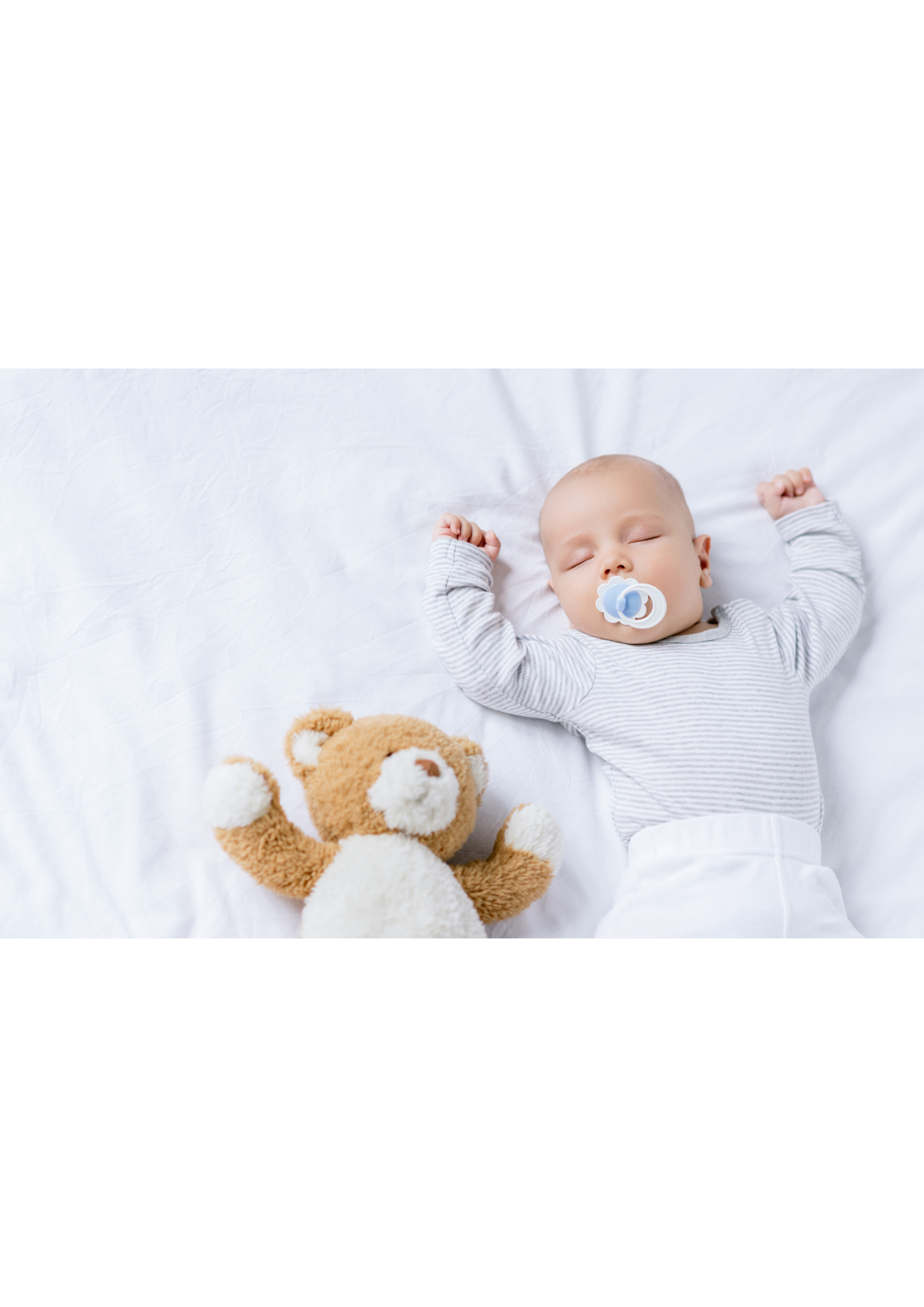 A sleeping baby with a pacifier in their mouth lies on a white bed with arms raised, next to a brown and white teddy bear.