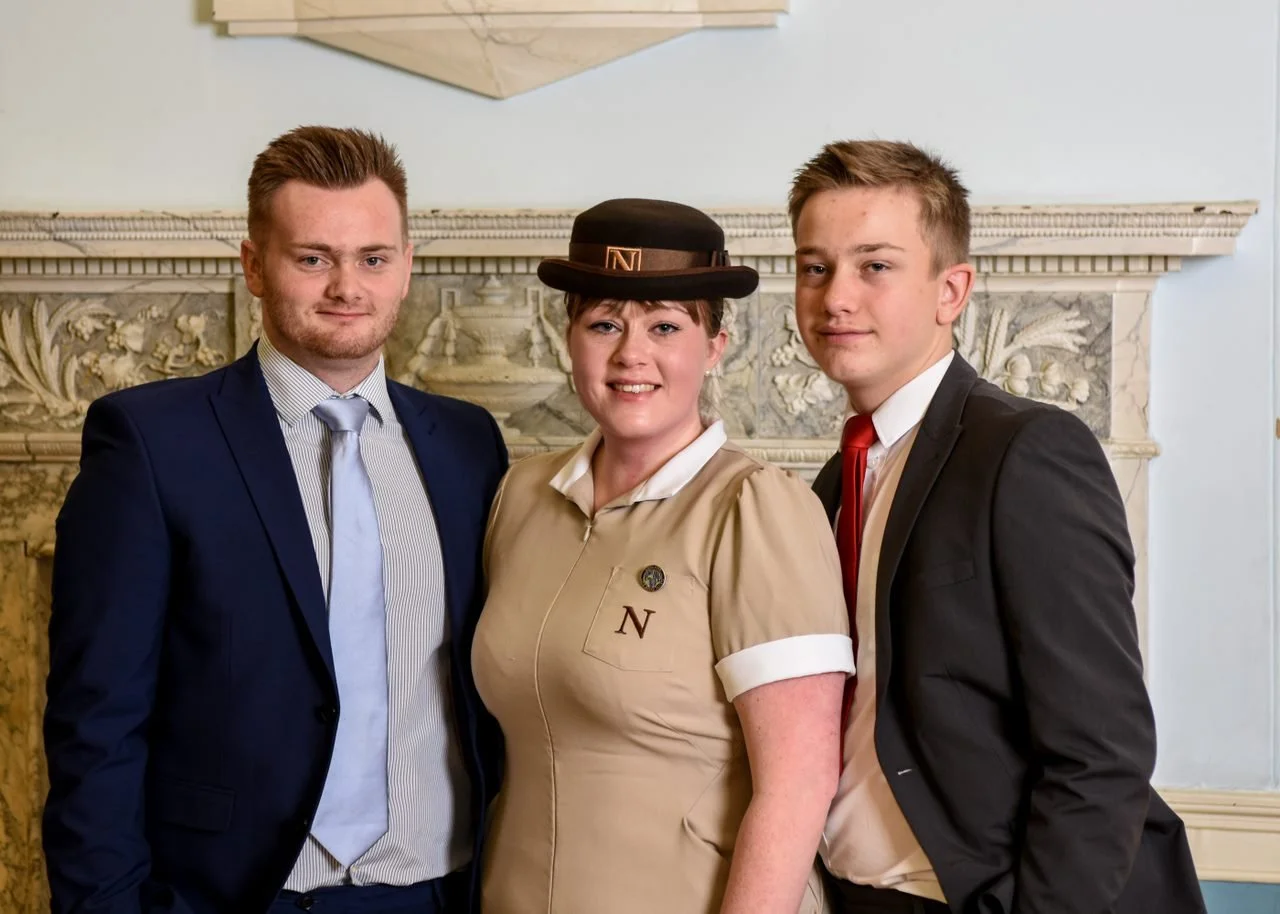 Three people in formal and costume attire standing in front of ornate fireplace, smiling at the camera.