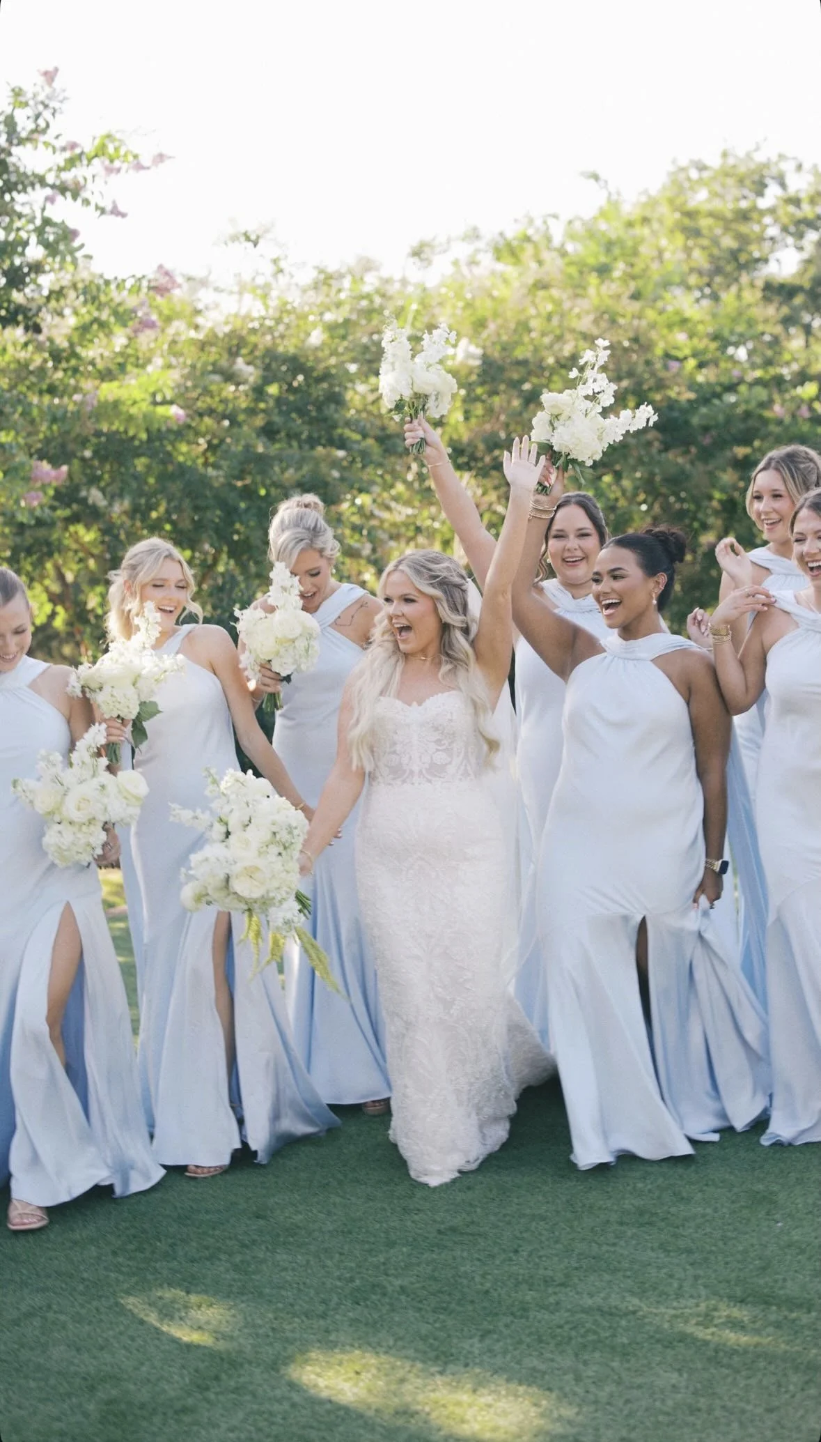 Bride in a white lace wedding dress walking with her bridesmaids dressed in matching white gowns, holding bouquets, celebrating outdoors on a sunny day.