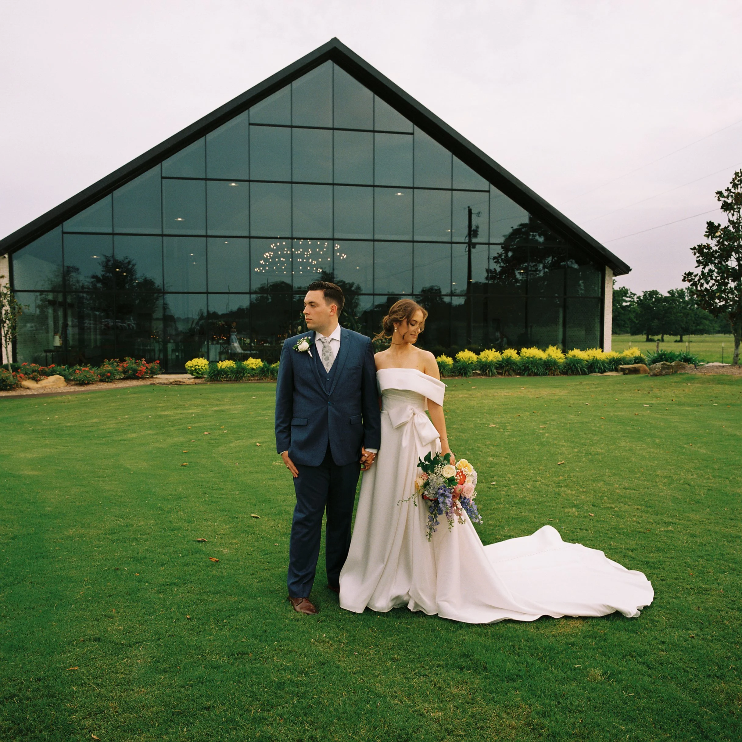 A bride and groom holding hands outdoors on a wedding day in front of a modern glass building with a lawn and flower bed behind them.