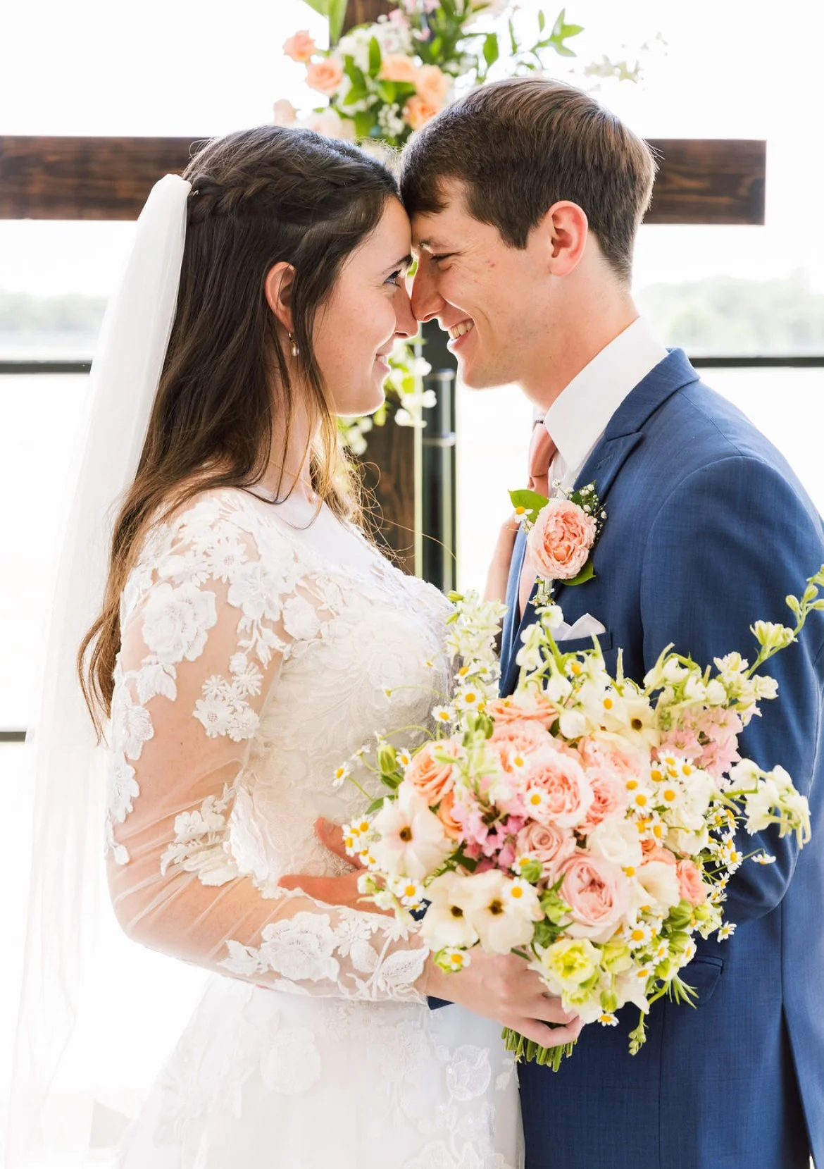 A bride and groom stand close during their wedding, touching foreheads and smiling at each other, with the bride holding a large bouquet of pastel-colored flowers.