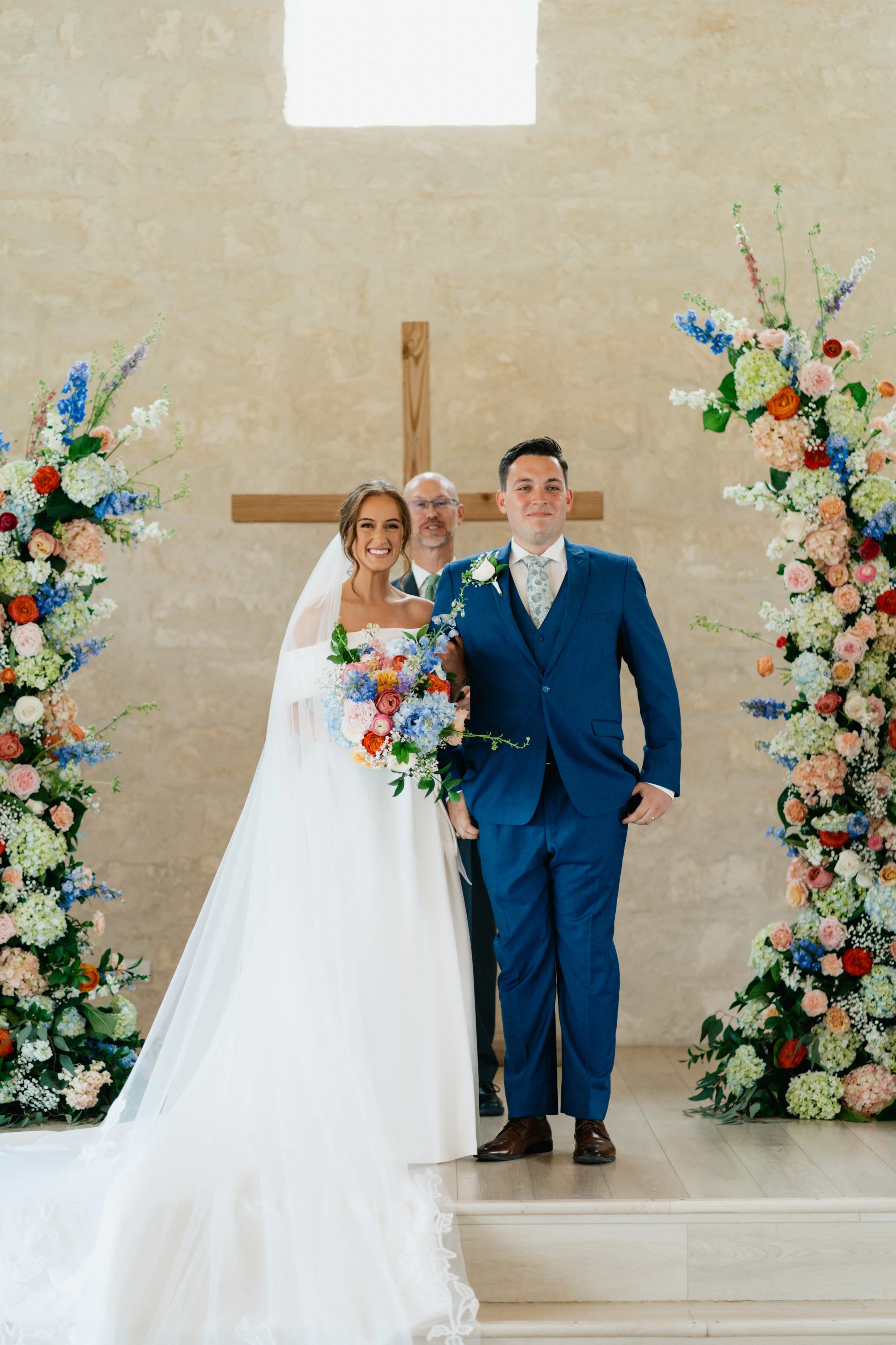 A bride and groom standing at the altar during a wedding ceremony, with a priest behind them, floral arrangements on either side, and a cross in the background.