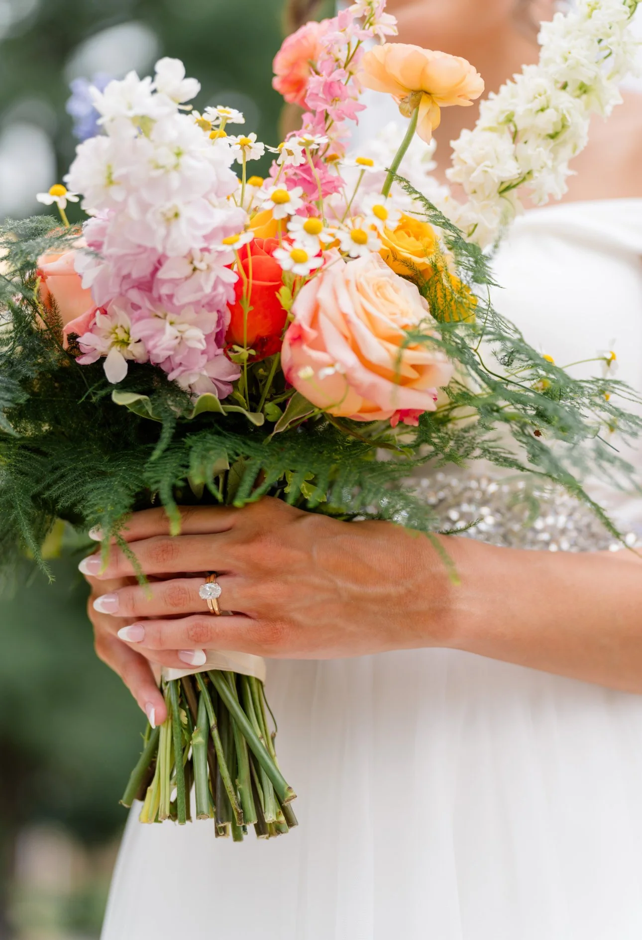 Close-up of a woman holding a colorful bouquet of flowers, including pink, orange, and white roses, daisies, and greenery. The woman is wearing a white dress and a ring on her left hand.