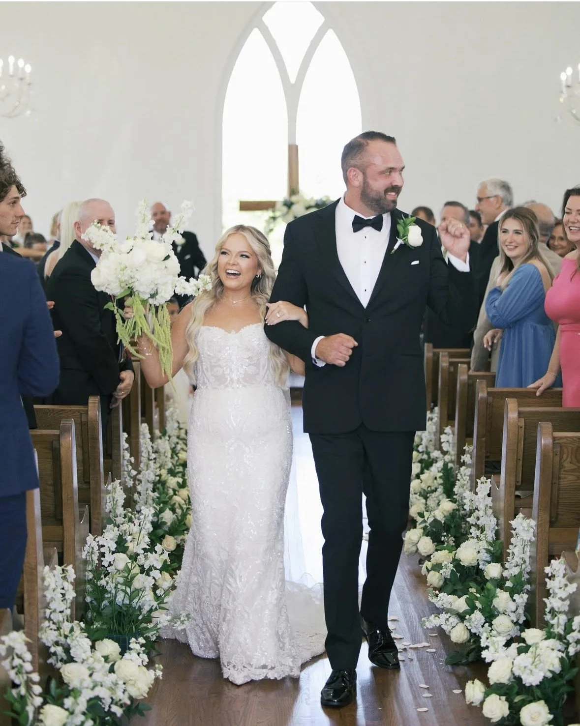 Bride and groom walking down the aisle at their wedding in a church, surrounded by guests and floral decorations.
