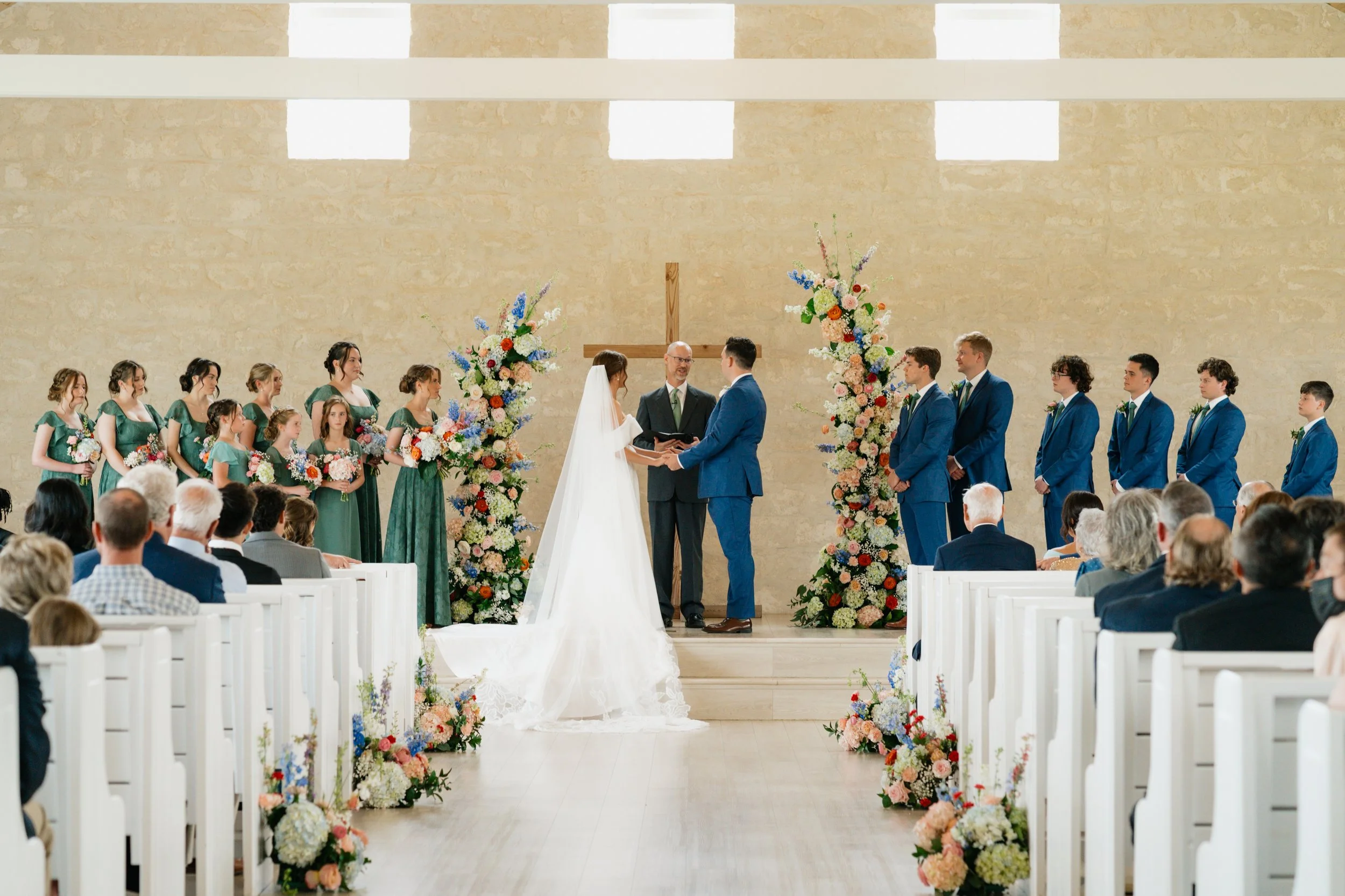 A wedding ceremony inside a church with a bride and groom exchanging vows, surrounded by bridesmaids and groomsmen, with decorated floral arrangements and guests seated in pews.