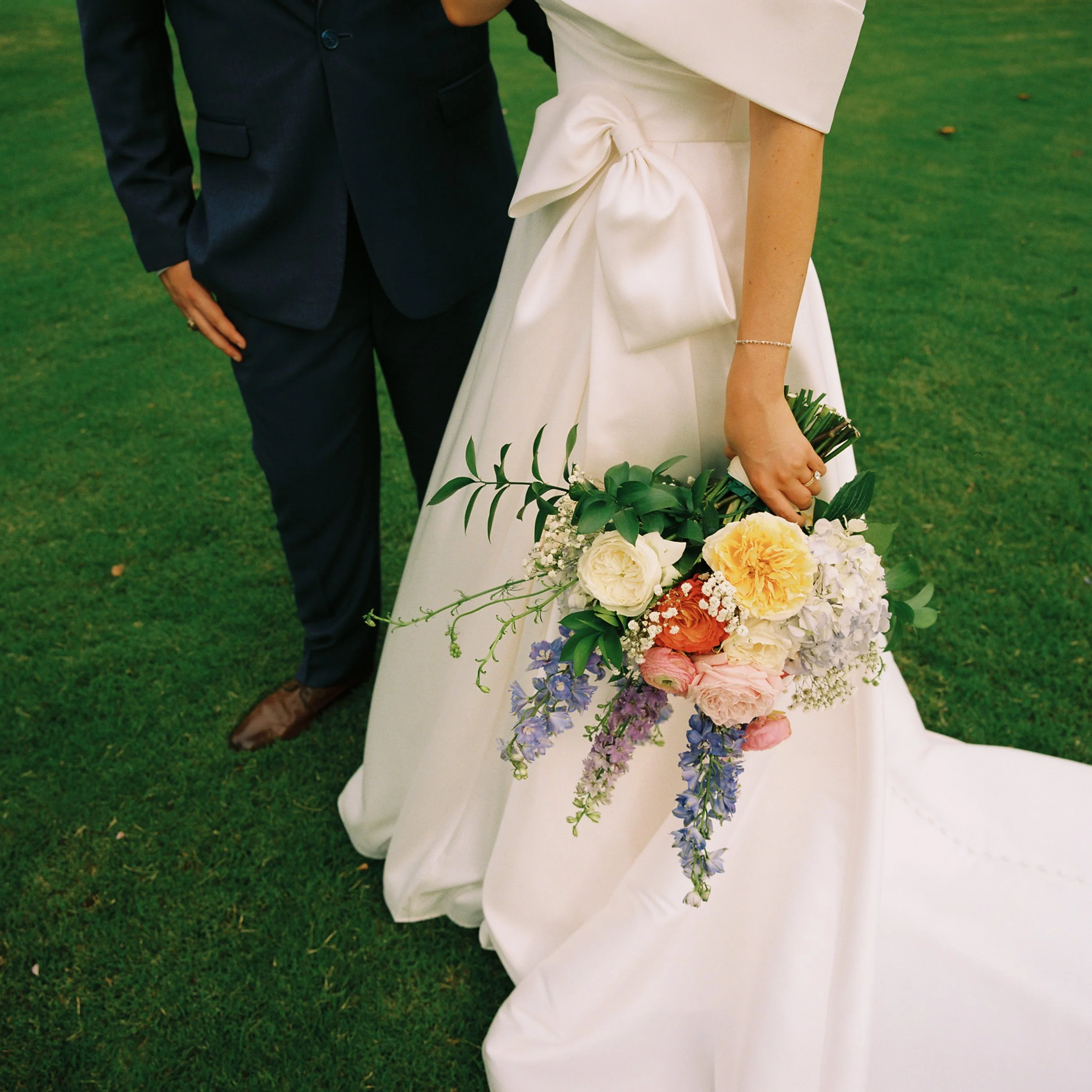Partially visible bride in white dress holding a colorful bouquet, standing next to groom in a dark suit on green grass.